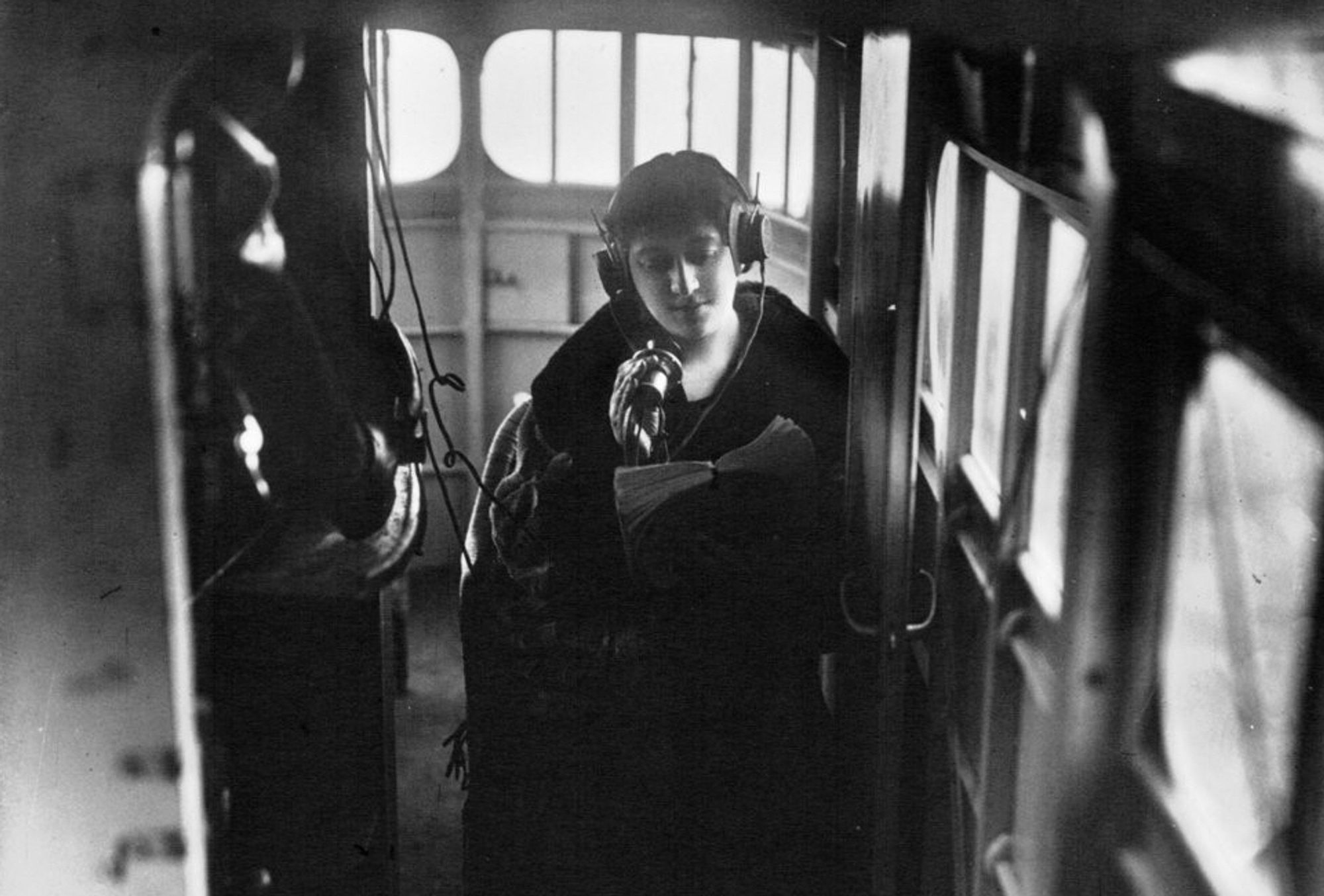 black and white photo taken inside the Goliath. the shot is lit by the front curved windows at the back of the shot (which are easy to mistake for the cockpit when viewed outside, but as with many aircraft of this era, the crew were sat on top of the fuselage, towards the wings, more exposed to the elements. a young woman wearing a coat is sat backwards and against the side of the aircraft, facing the camera. she is wearing chunky headphones and looking down at a book she is holding with one hand. she is holding a microphone in the other, and both this and the headphones are wired to some communications equipment on a compartment wall next to her, that are not properly visible in the dimly lit shot