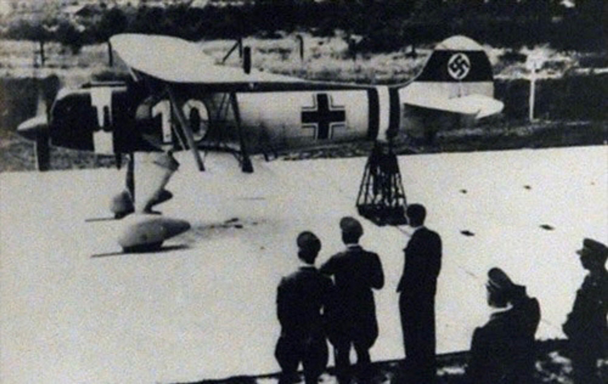 black and white photo of a Heineken He 51 fixed horizontally on the ground via its tailskid, engine running. officers in uniform are studying the aircraft, their backs to the camera, alongside a dark-suited man with his hands in his pockets