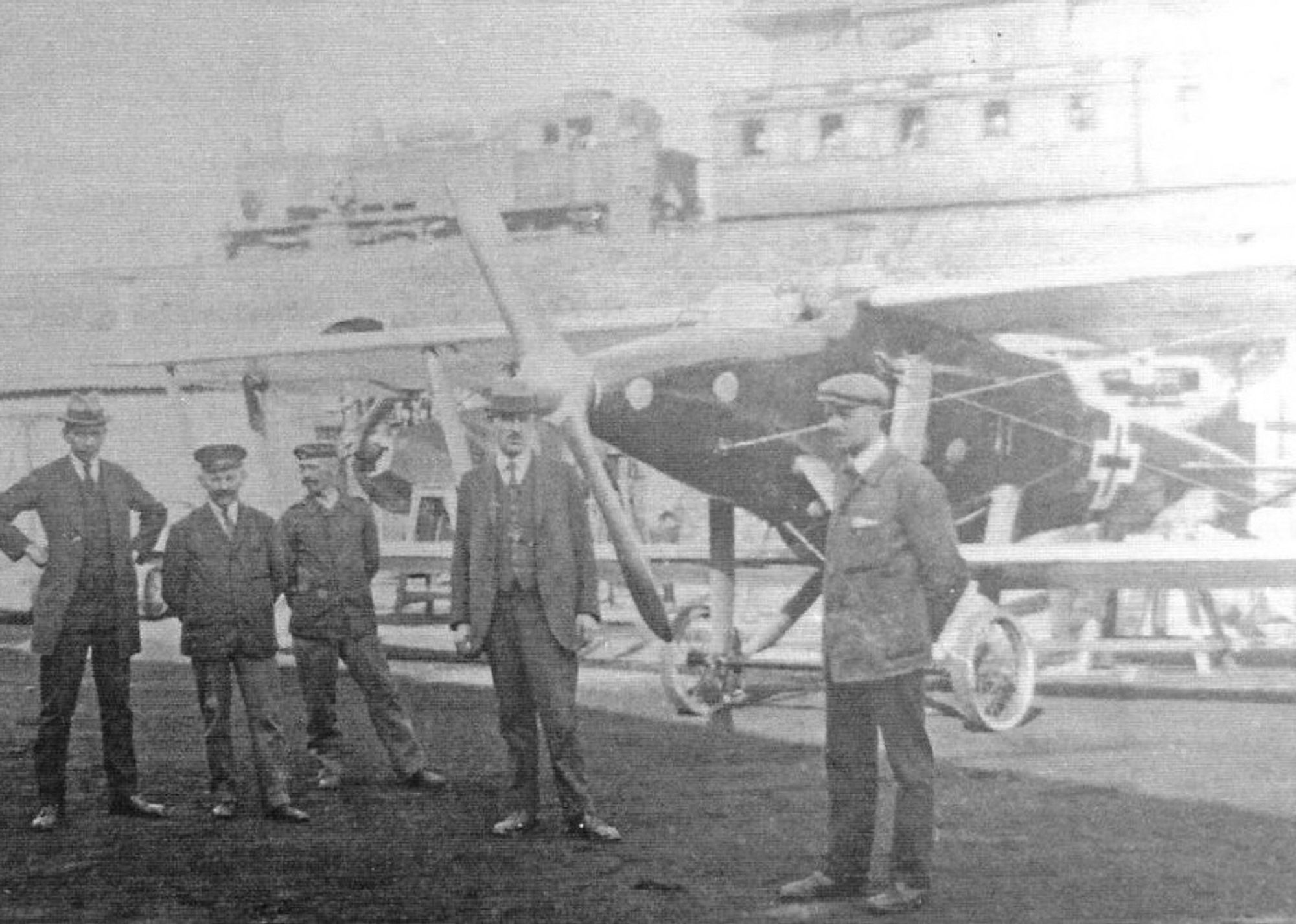 black and white photo of the aircraft in front of a low railway bridge, while a steam train with passenger carriages passes closely by (we can just about make out people in the windows). the aircraft is a large, thin streamlined biplane, with relatively long looking wings. the open cockpit with front windscreen is situated directly in front of the top wing, that appears to be fixed to the top fuselage. the most striking feature is the long, tapered rattish nose, ending in a large propellor that looks glued on. in the foreground, several smartly dressed men in an array of caps and hats pull a range of poses