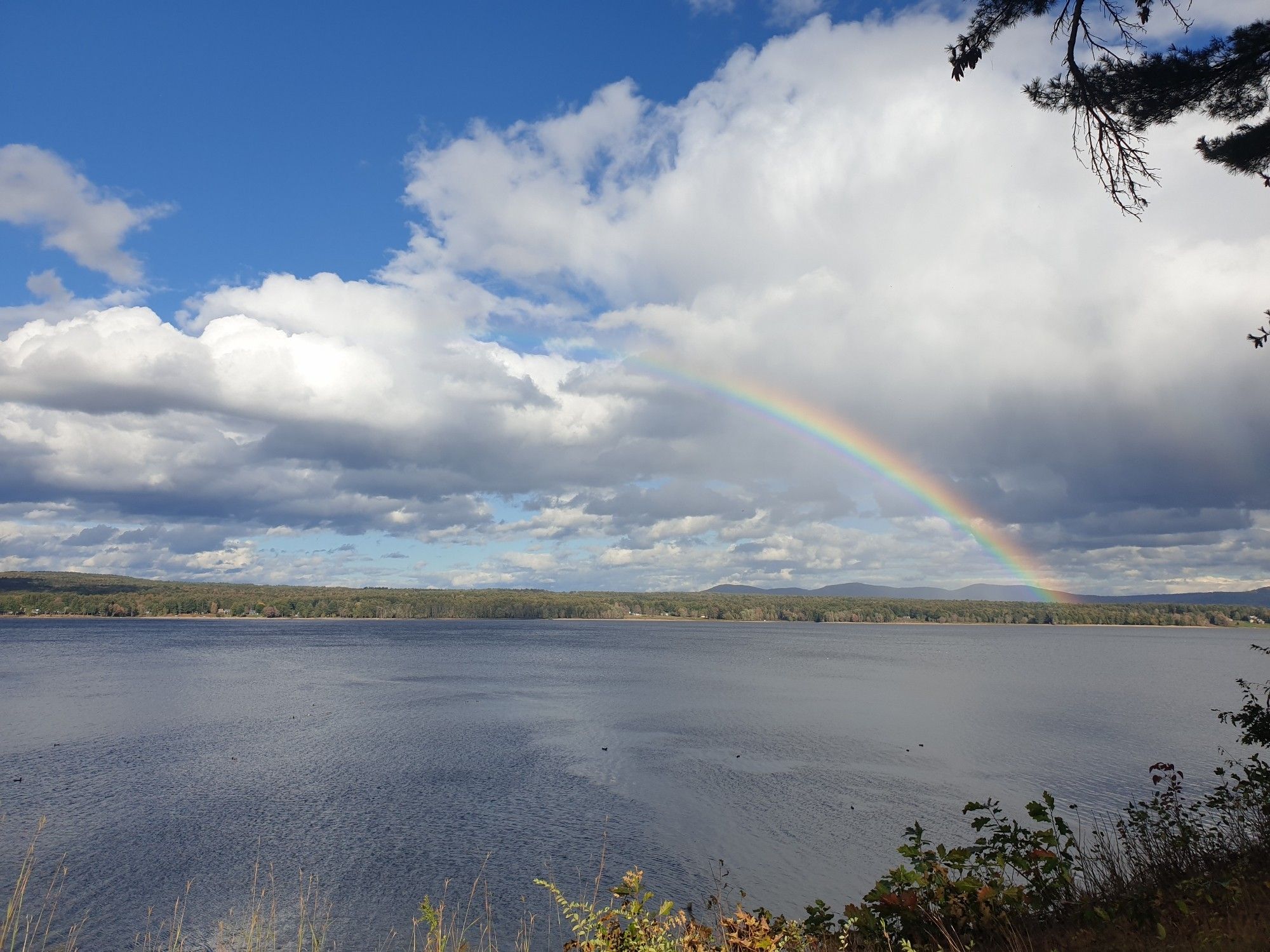 A large lake with a partly cloudy but bright blue sky, with a rainbow on the right connecting the clouds to the ground.