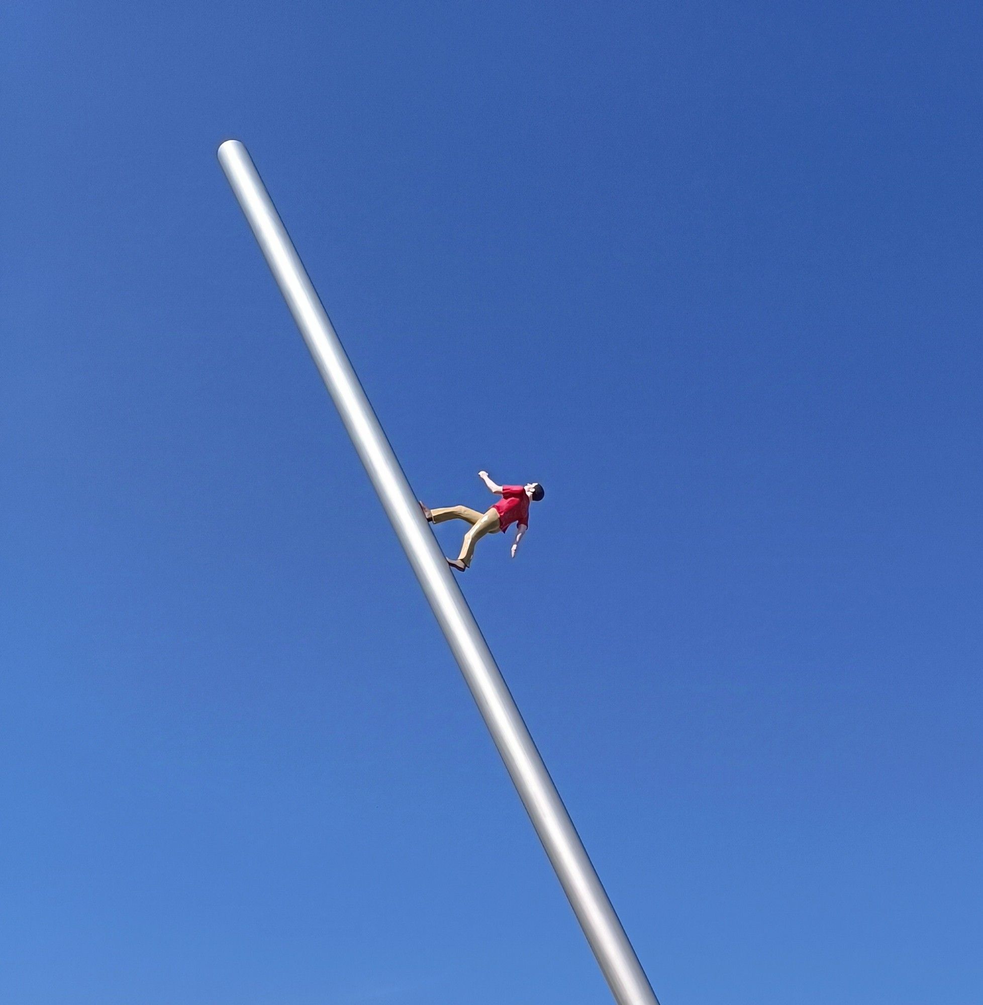 Der Himmelsstürmer in Kassel. Wolkenloser blauer Himmel, die Figur eines Mannes mit heller Hose und rotem Shirt, die an der Stange entlang Richtung Himmel läuft.