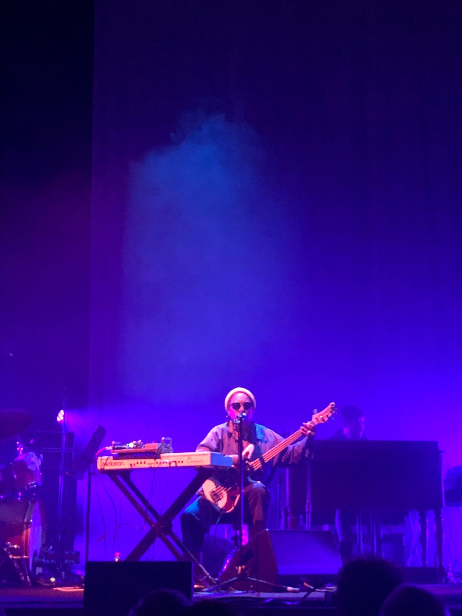 A woman sitting behind a keyboard, playing a bass guitar, singing into a mic on a stand, bathed in atmospheric blue and purple light.