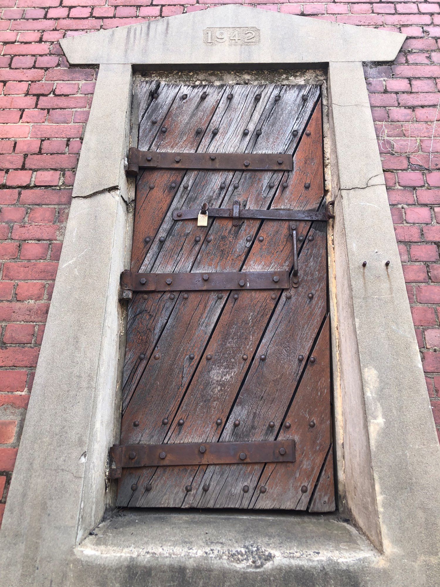 Large, old wooden door in a cement frame stamped 1942 with hefty iron hinges, nail heads handle and padlock.