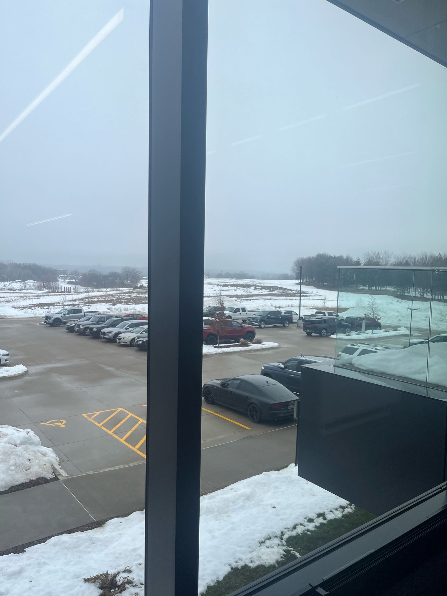 View out a window over a parking lot to snow covered fields and fog covered city in the distance.