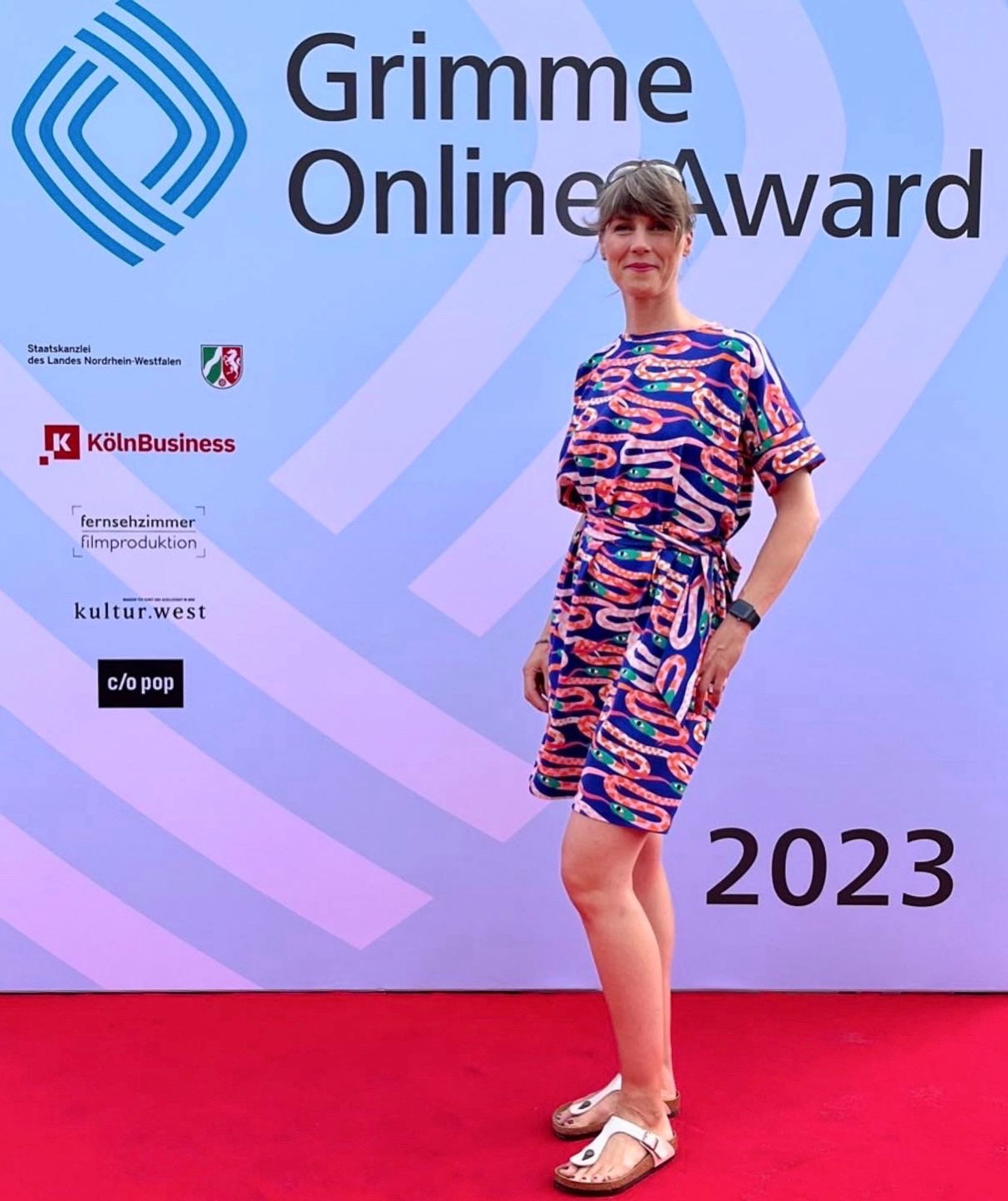 Nora Hespers steht auf dem roten Teppich vor der Fotowand, auf der deutlich Grimme Online Award zu lesen steht, sowie das Jahr 2023 und einige Sponsoren. Sie trägt ein schickes blaues Kleid mit einem grafischen Muster mit verschiedenfarbigen Schlangen in Rottünen in der Form eines plattgedrückten S.