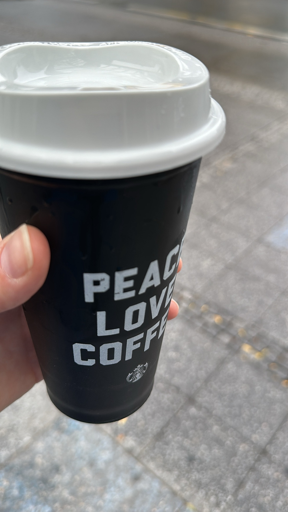 A hand holding a black reusable cup with the words peace love coffee in white printed on it