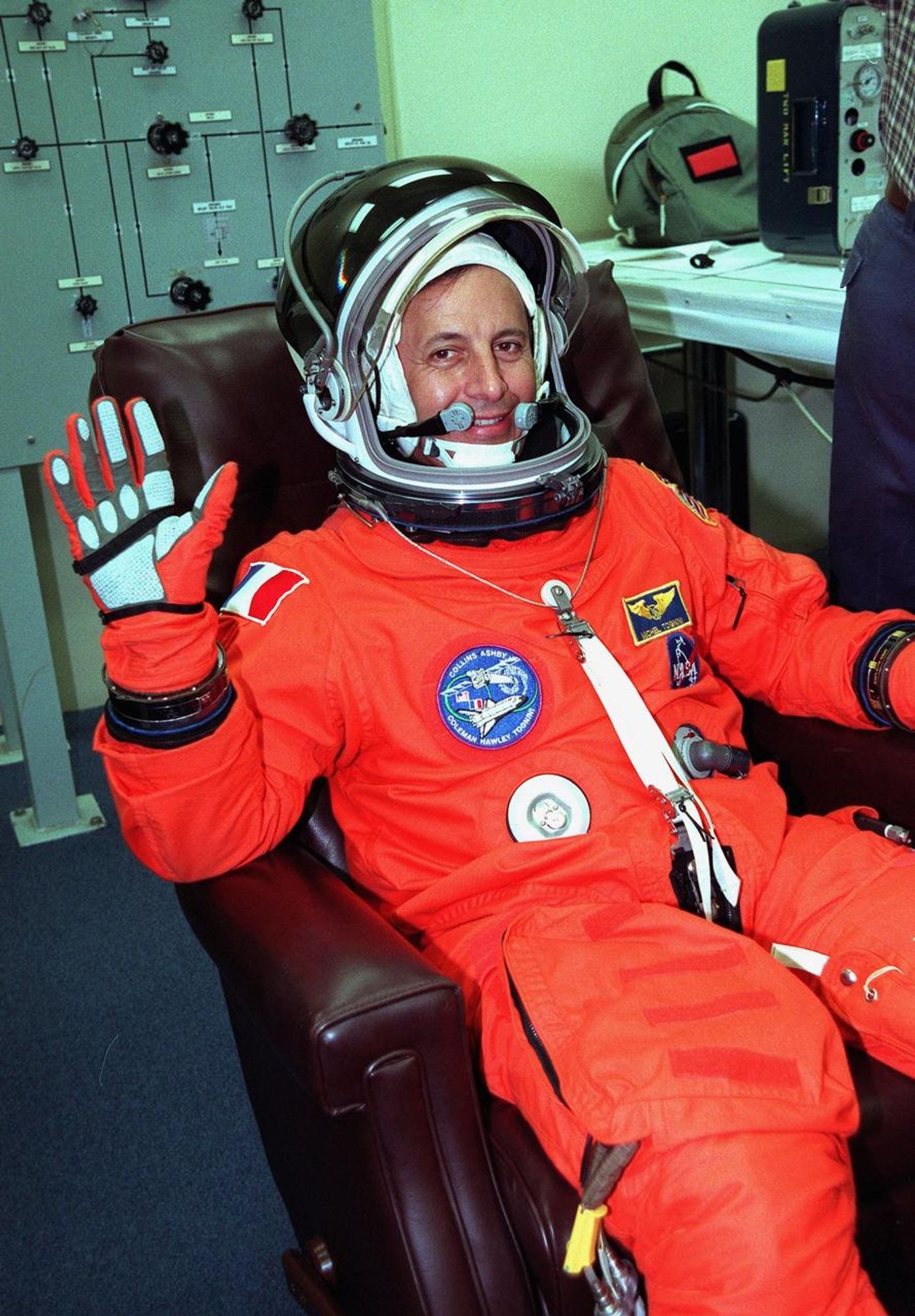 Michel Tognini, wearing his bright orange launch and entry suit, waves at the camera and smiles.