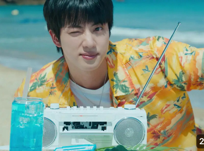 Screen grab of Jin in Supertuna MV. Jin is winking cutely on the beach wearing a yellow Hawaiian print shirt and white t shirt.