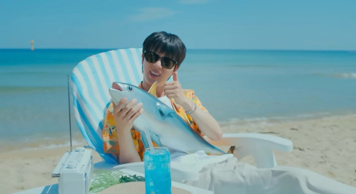 Jin giving thumbs up holding a super tuna for his Supertuna MV He is sitting on a blue and white lounge chair at the beach wearing yellow Hawaiian shirt