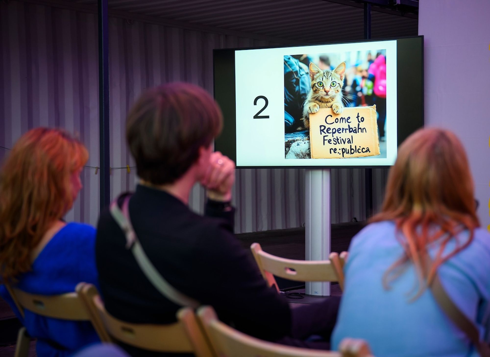 Zuschauende während des Prompt Battles. Auf einem Bildschirm ist eine Katze mit Schild zu sehen, auf dem "Come to Reperrbahn Festival re:publica" steht