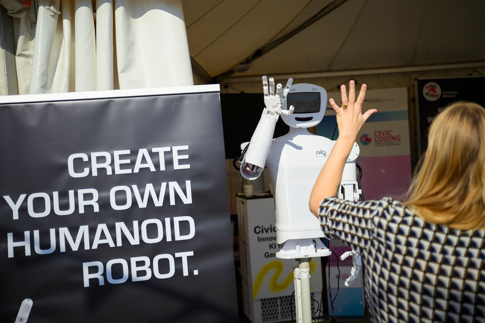 "Create your own humanoid robot" auf einem Schild neben einem humanoiden Roboter, der einer Besucherin High Five gibt