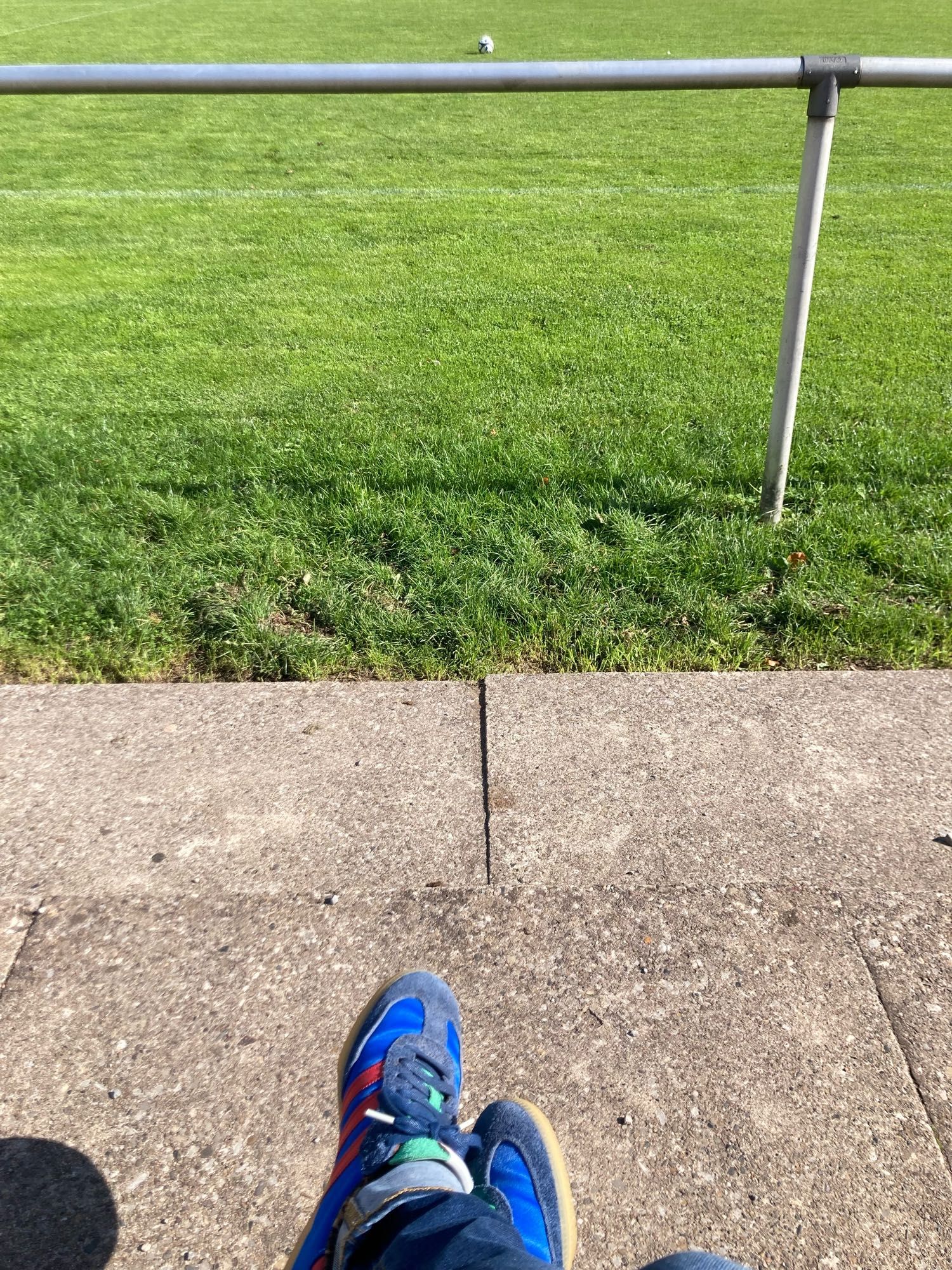 Schicke Schuhe vor Betonstufen mit Blick auf eine Sportplatzumrandung aus Stahlrohrund sattem Rasen dahinter. Ein Fussball liegt auf dem Rasenm