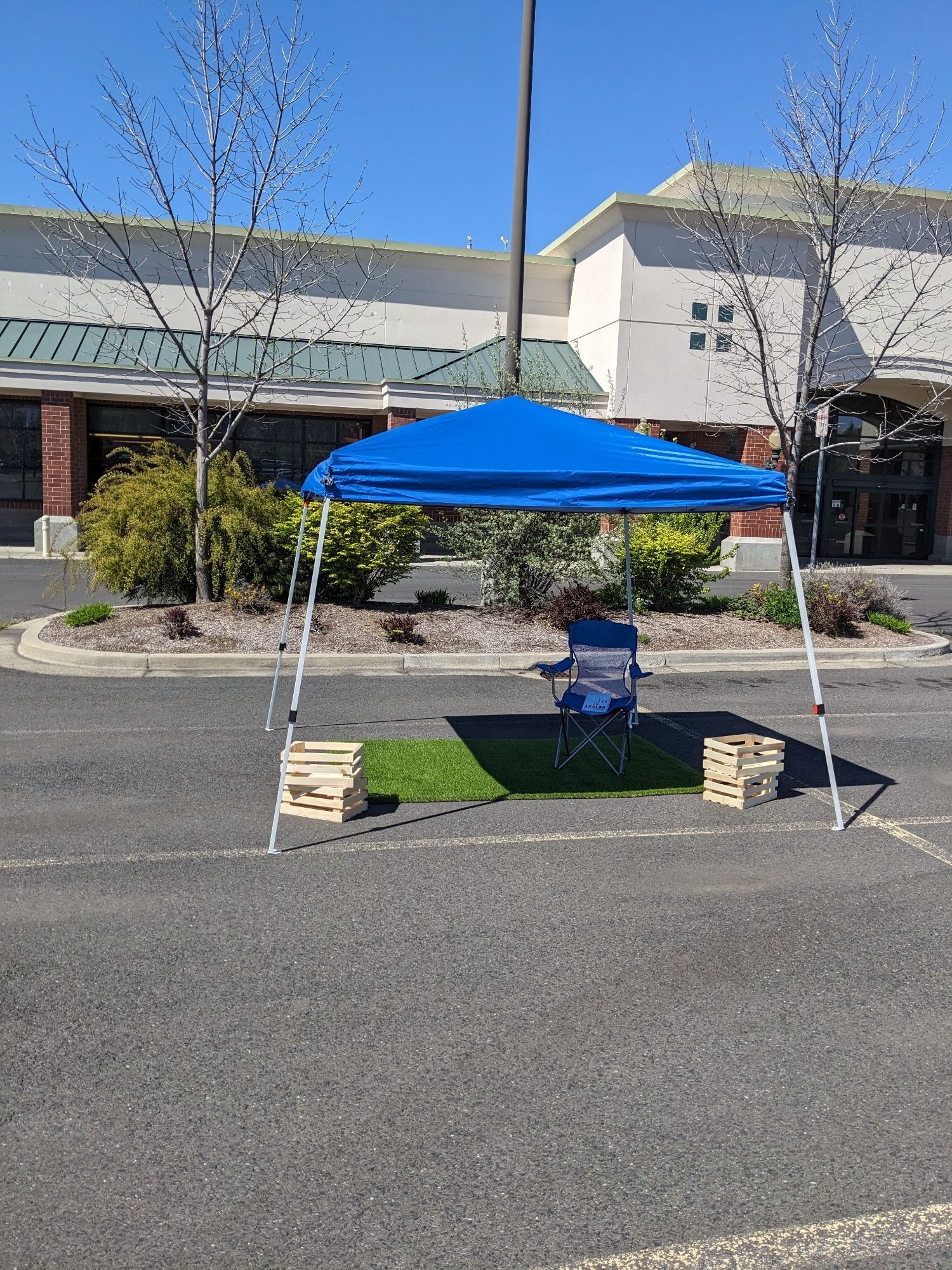 A blue temporary canopy over a patch of artificial turf in an empty parking lot. There are small wood crates on the corners of the canopy, and a lawn chair in the shade provided.

On the chair is the book "The Future is Analog" by David Sax