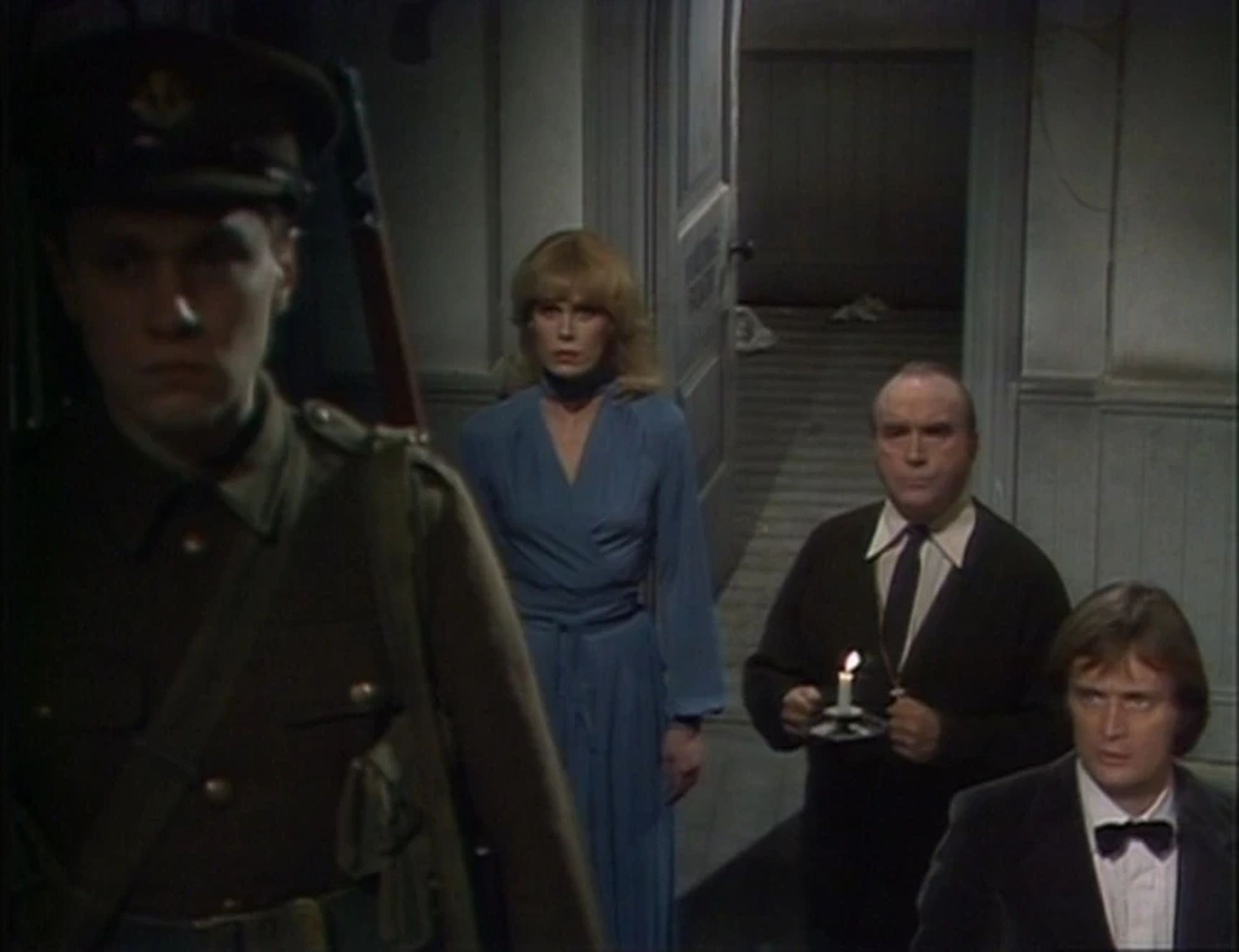 Three people stand at the foot of a staircase ins a dilapidated building, looking up to a fourth person, a World War 1 soldier, who is standing at the top. The Soldier is facing away from the others, looking towards the camera. 

The three standing at the bottom from left to right are Sapphire, played by Joanna Lumley, a tall, young, blonde haired white woman in a glamorous blue dress, Tully, played by Gerald James, a stout, middle aged white man wearing a cardigan, shirt and tie, holding a candle, and lastly Steel, played by David McCallum, a young white man with longish 1970s style light coloured hair, wearing a black dinner suit, white shirt, and black bow tie.