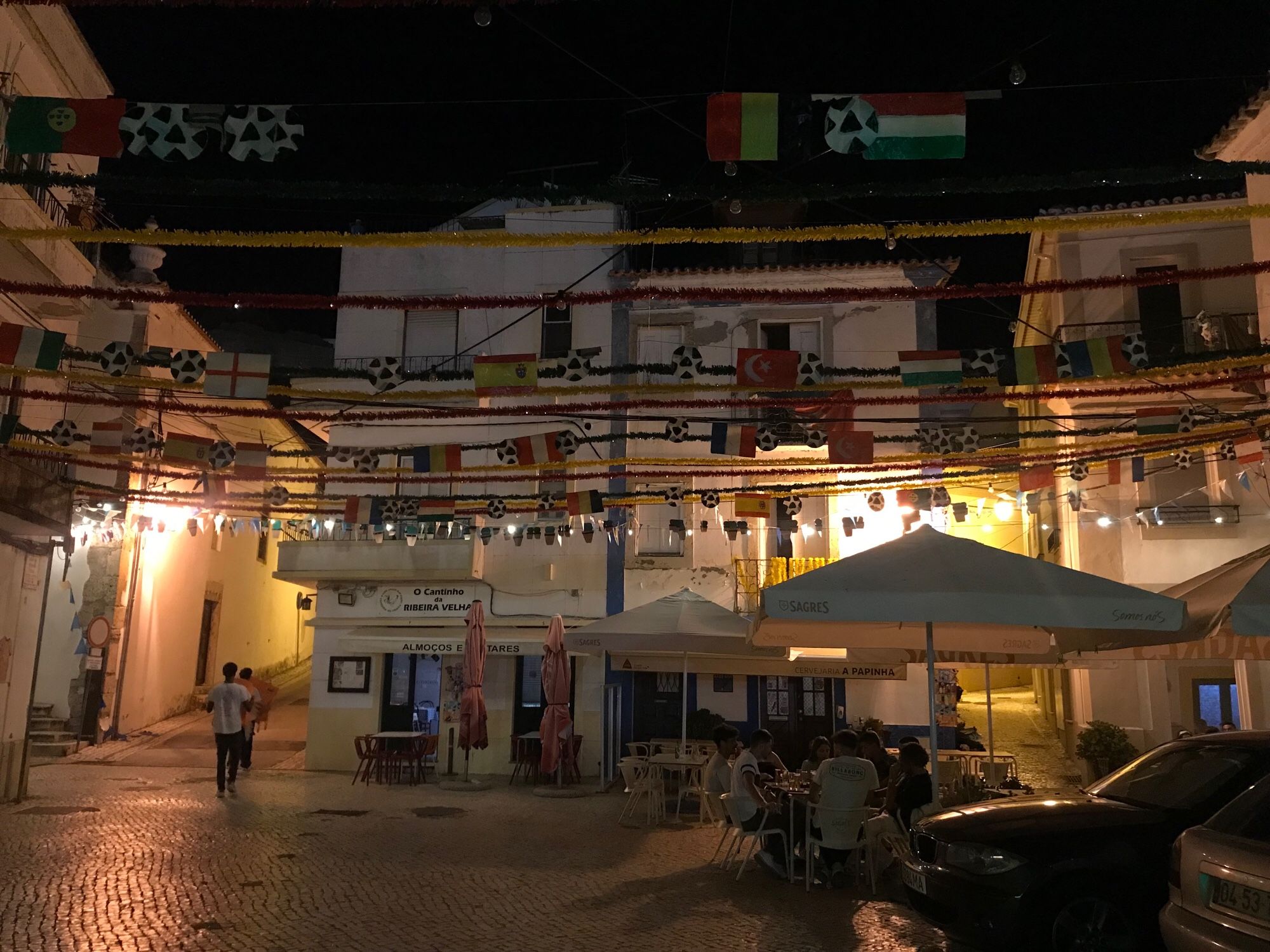 Portugal. Blick auf einen kleinen Platz mit Restaurant und Bar, in der Nacht, mit Beleuchtung. Es sitzen noch Gäste an Tischen. Es hängt noch die Dekoration von der EM an Bändern in der Luft.