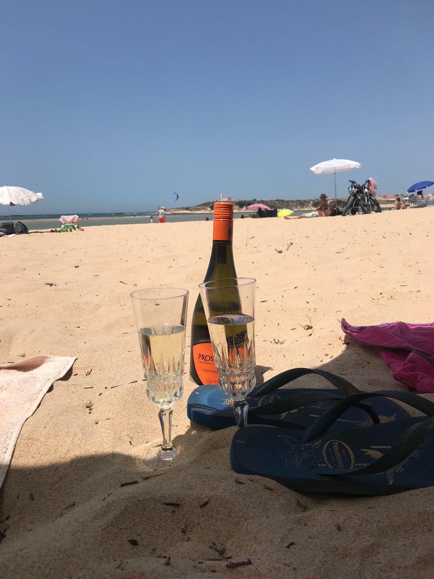 Eine Flasche Prosecco mit zwei gefüllten Sektkelchen, im Sand, am Strand, mit Blick Richtung Meer. Ein blaues Paar FlipFlops vollenden das Stilleben.