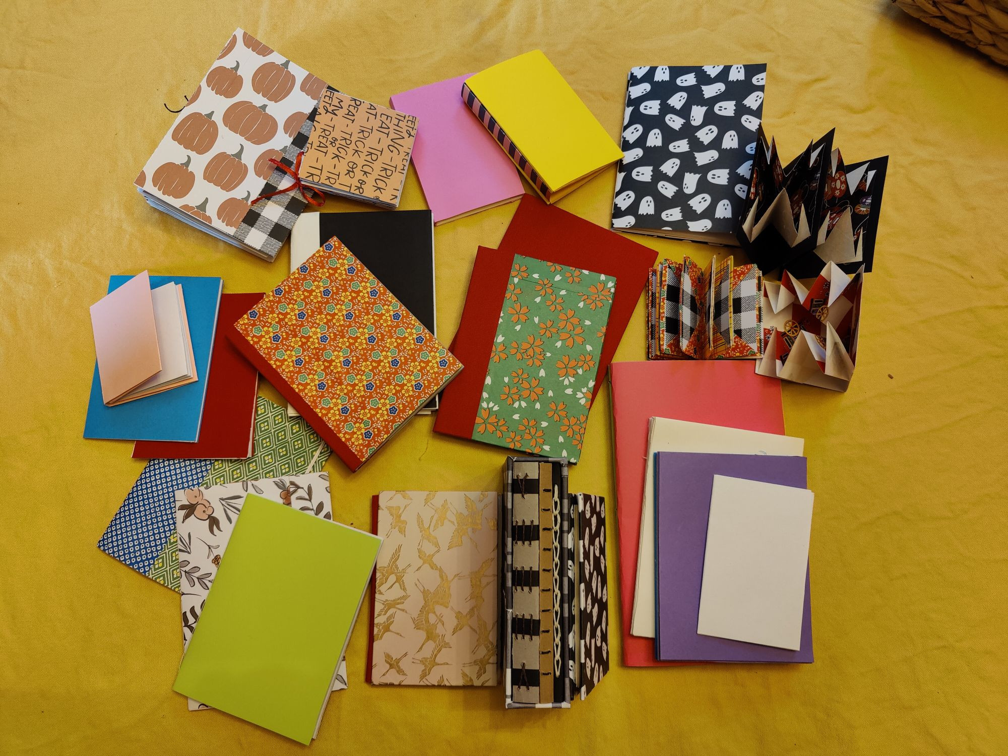 image of lots of little blank books in different colors scattered across a yellow table. there's tons of halloween paper and origami paper used for covers.