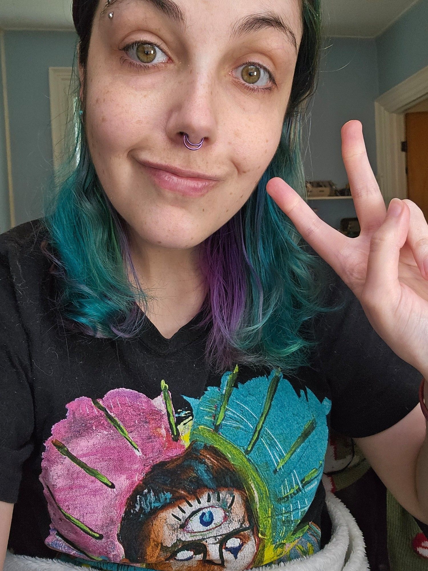 Rae, a white nonbinary person with shoulder length purple and turquoise hair, hold up her fingers in a peace sign. Rae is wearing a bright Of Monsters and Men t shirt with a man with three eyes surrounded by yellow sunbeams over a pink and blue background