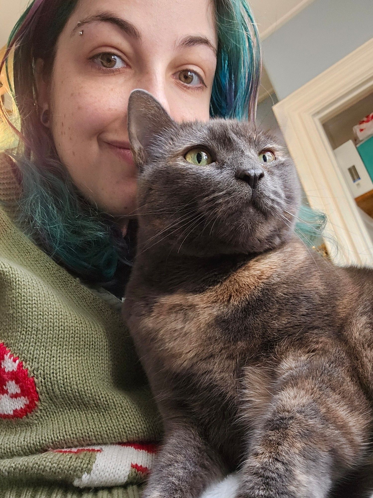 Rae, a white nonbinary person with turquoise and purple hair, smiles at the camera. On her lap is Isis the dilute tortie cat.