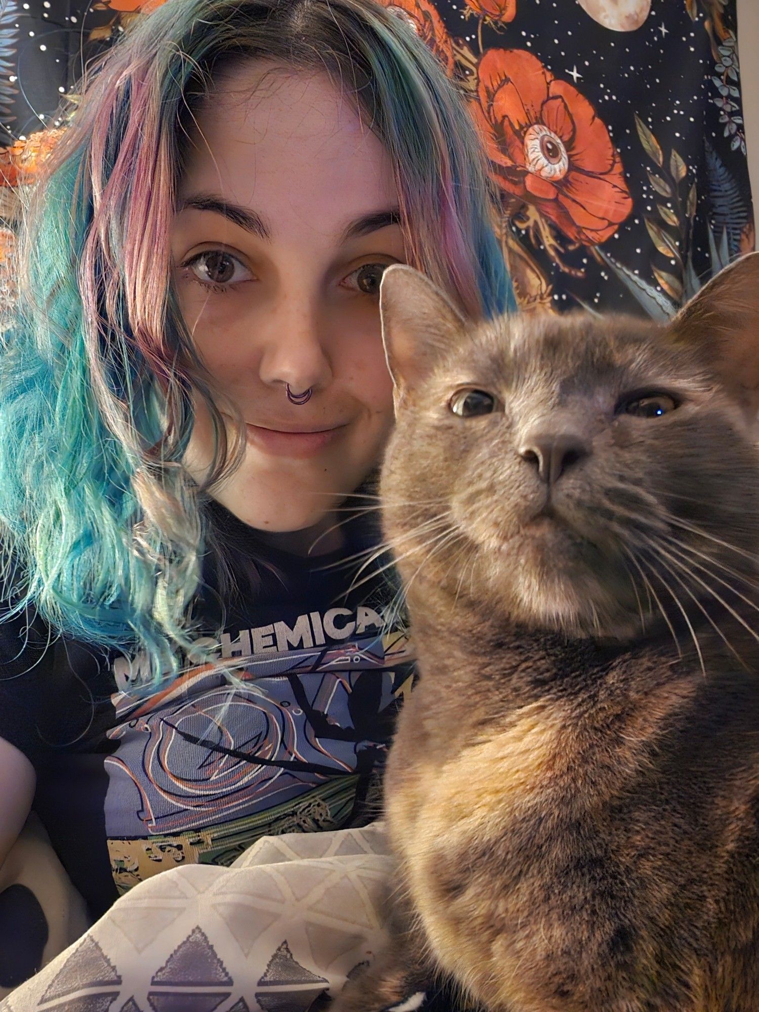 Rae, a white nonbinary person, smiles at the camera. They have shoulder length turquoise and purple hair. On their lap is Isis the dilute tortie cat. Isis looks to the camera with a silly face