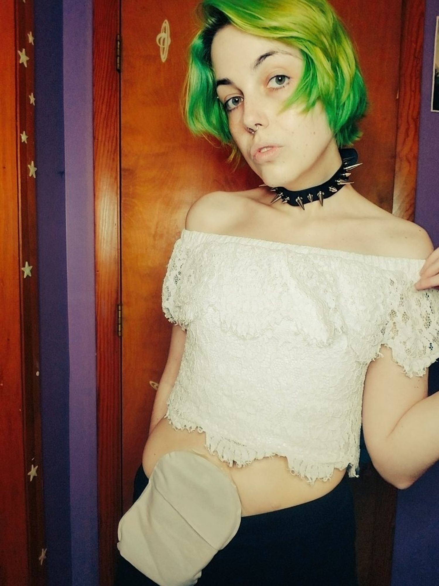 Rae, a white nonbinary person with green hair, poses for the camera. They are wearing a lacy white crop top and a spiked black choker. Rae has her ostomy bag visible in the photo!