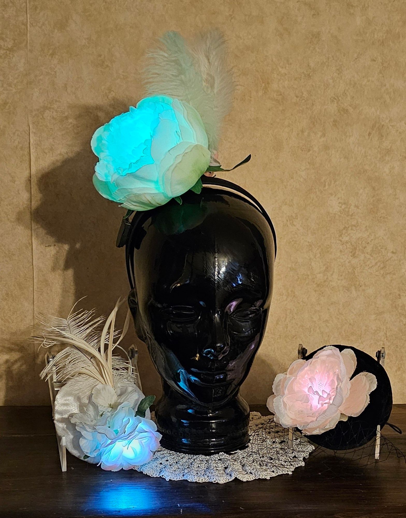 Three fascinator hats with color-changing LEDs. In the center is a large white flower attached to a headband displayed on a vintage black glass display head. The LED is glowing blue. On the right is an all-white fascinator with a flower and feathers. The LED is glowing blue. On the left is a black velvet cocktail hat with a white flower and black veiling. The LED is glowing red.