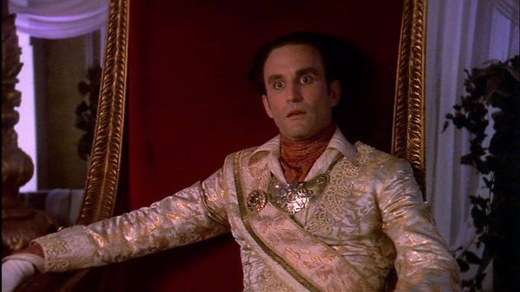 Emperor Cartagia, a man wearing an ostentatious coat in white and gold as well as a large golden medallion, sitting on a golden throne with red upholstery, looking surprised.