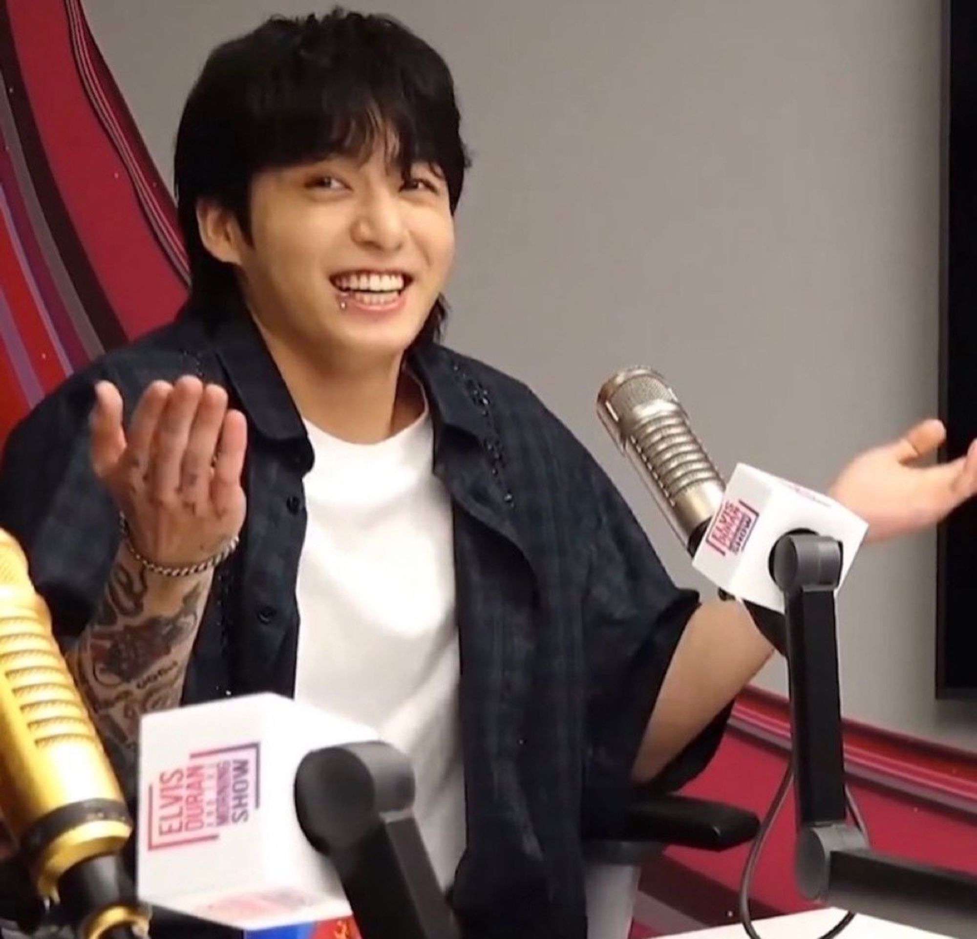 Jungkook of BTS being interviewed by a radio station, he has a wide smile and mischievous eyes his arms are doing an i don't know gesture