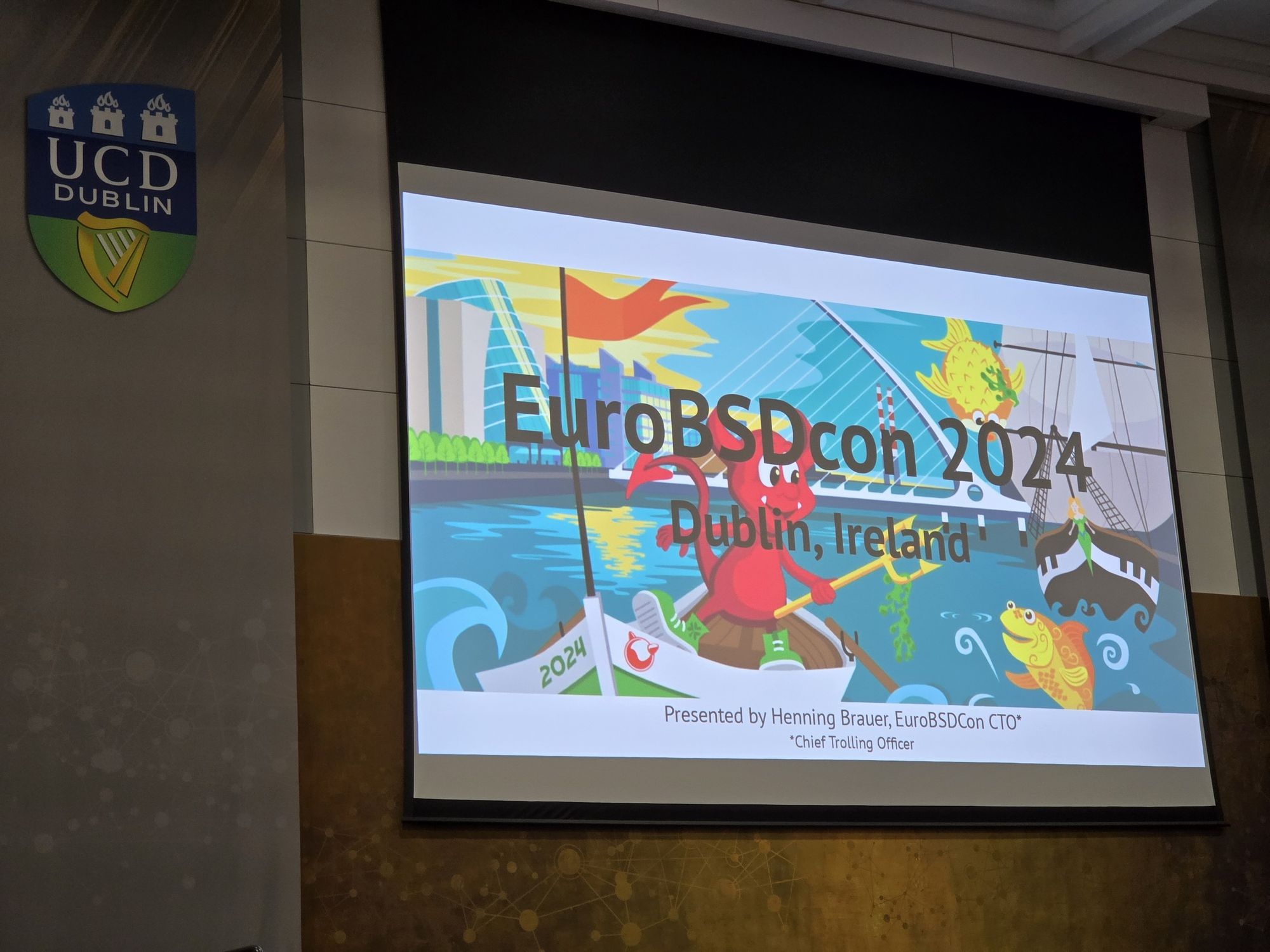 A projected slide from the EuroBSDcon 2024 event in Dublin, Ireland. The slide features colorful artwork of a red BSD daemon paddling a small boat in a river with a modern cityscape in the background, including a bridge, buildings, and a green sea monster. The UCD Dublin logo is visible on the left side of the image, signifying the conference location. The slide indicates that the presentation is being given by Henning Brauer, titled "EuroBSDcon CTO Chief Trolling Officer".