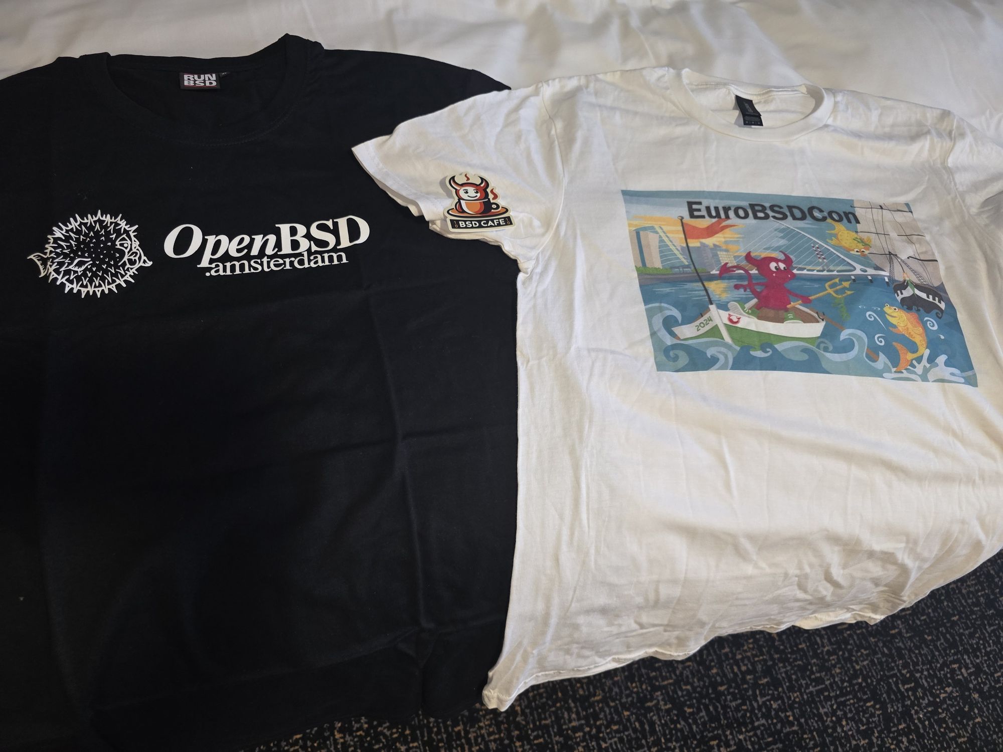 The image shows two t-shirts side by side along with a "BSD Cafe" sticker. The black t-shirt on the left features the text "OpenBSD.amsterdam" in white, alongside the iconic OpenBSD pufferfish logo. The white t-shirt on the right has a colorful design for EuroBSDCon, displaying the event logo and a BSD daemon sailing a small boat in a whimsical sea with fish and a city skyline in the background. Near the top of the white t-shirt is a "BSD Cafe" sticker, which features a mascot and the words "BSD Cafe." This collection showcases some unique BSD-themed souvenirs from EuroBSDCon.