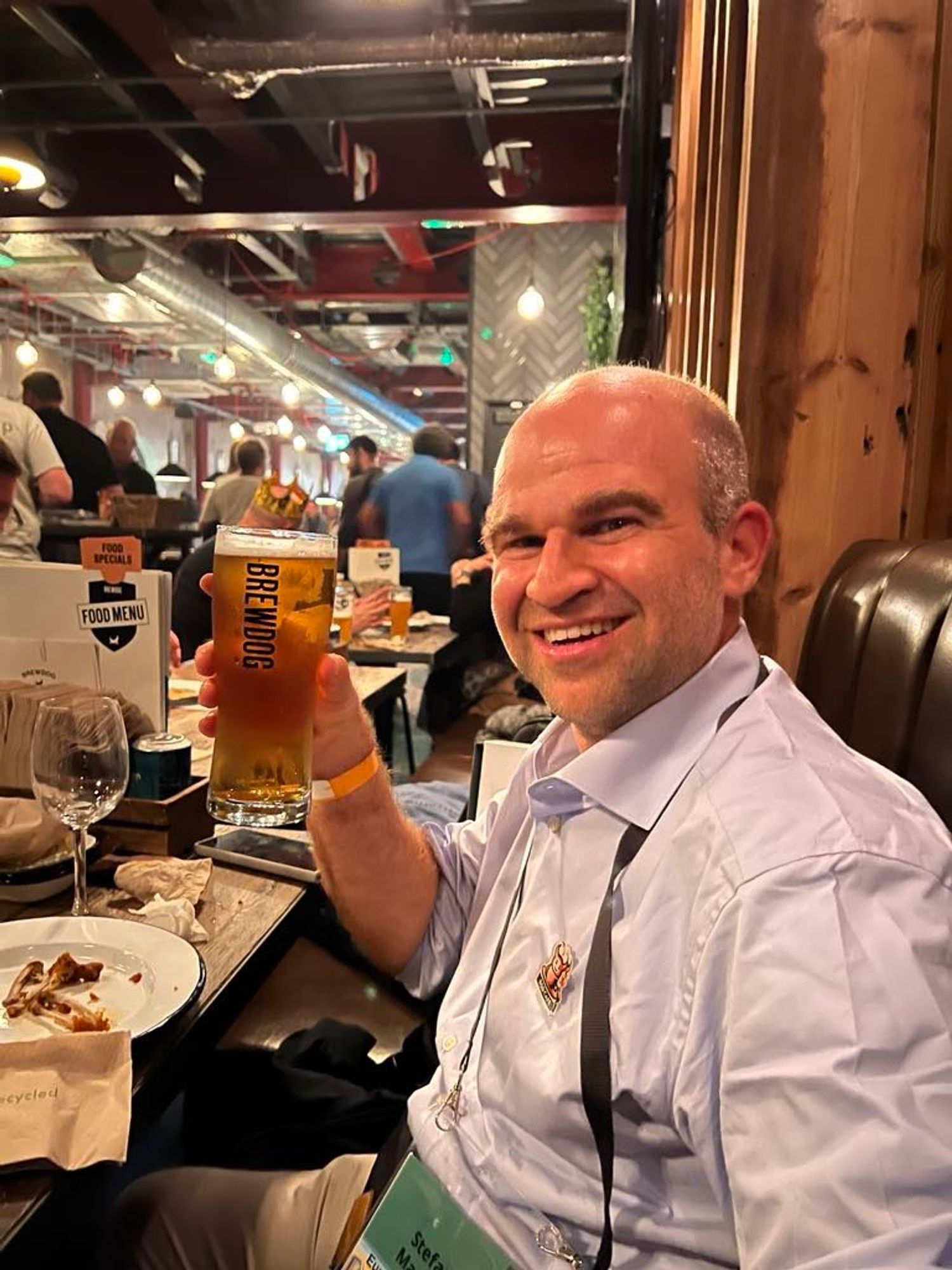 It's me, Stefano Marinelli, wearing a light blue shirt and sitting at a restaurant table, holding a glass of beer with the "BrewDog" logo. I am smiling at the camera, appearing to enjoy the EuroBSDCon social event, in Dublin. On my shirt, I am wearing a "BSD Café" pin. The background features other people at tables, with industrial-style ceiling fixtures and warm, ambient lighting. Plates, napkins, and a wine glass are visible on the table in front of him, adding to the casual, friendly atmosphere.