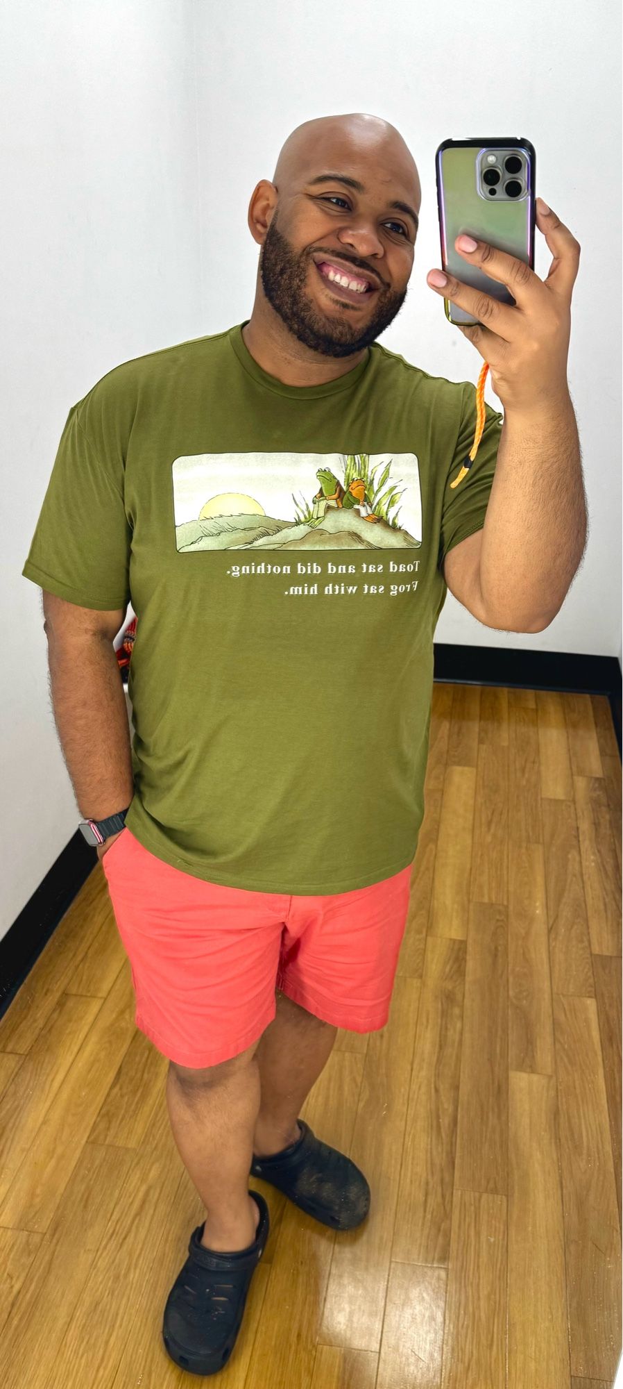 Guy wearing pink shorts and a green top with frog and Toad image on it in front of a mirror in a dressing room.