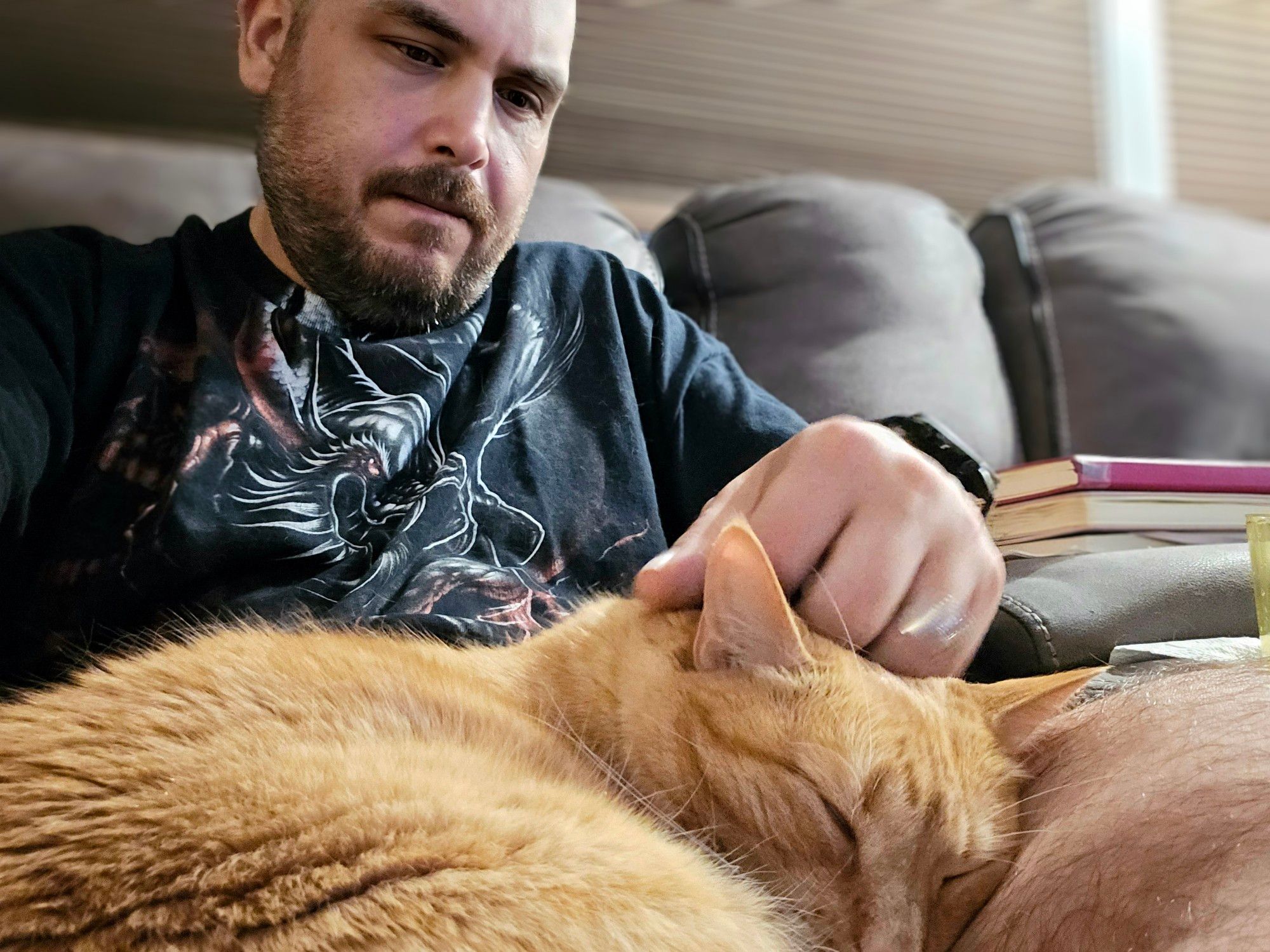 Matt B, wearing a dark graphic T-shirt, pets a sleeping orange cat on his lap. The scene appears relaxed, with books stacked on the couch beside him.