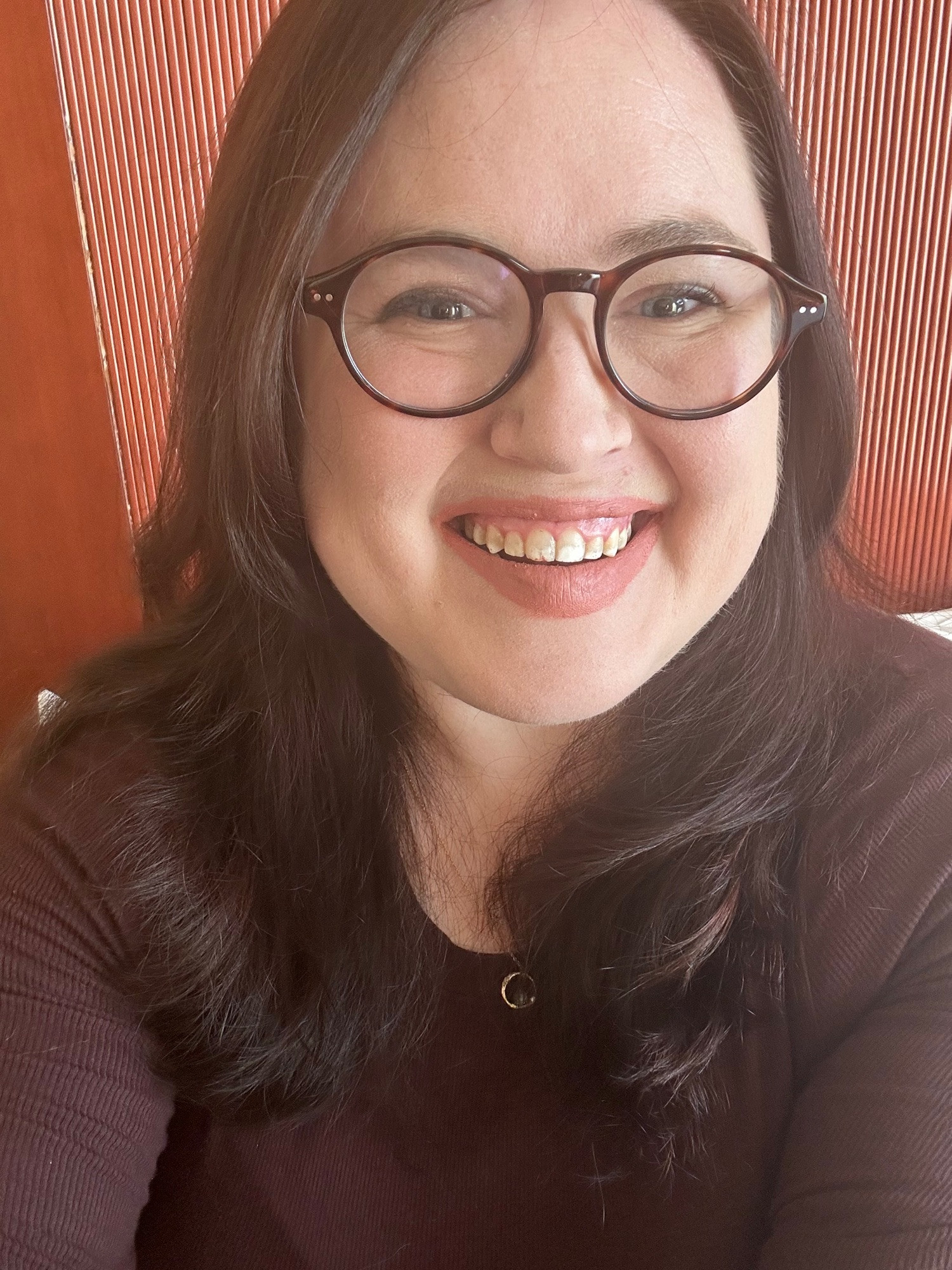 White woman with long brown hair and bangs smiles at the camera. She is wearing glasses and makeup with a long sleeve brown shirt.