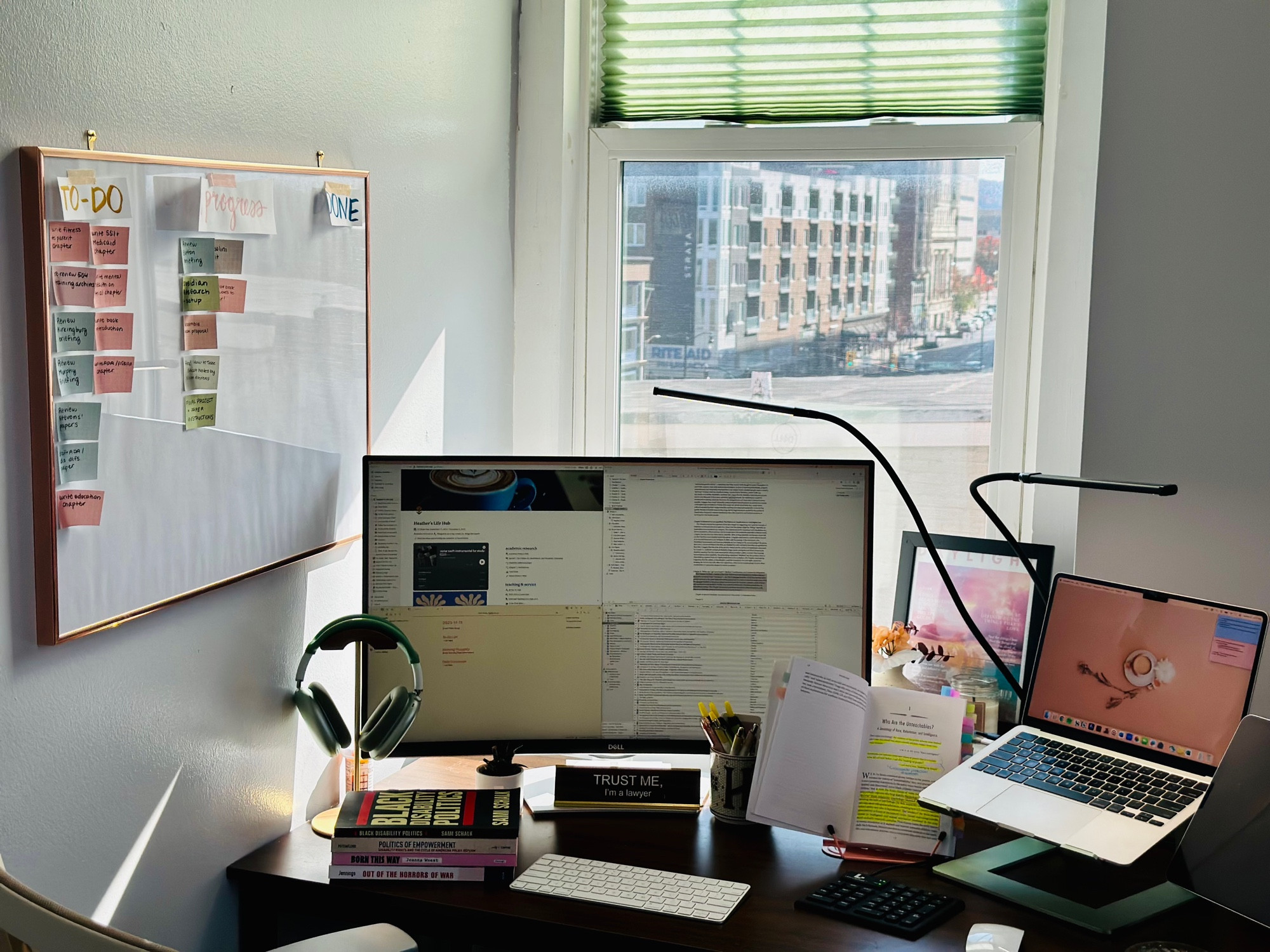 A desk set up with a laptop, an external monitor, several books, a few different types of keyboards, and a light lingering. There is also a kanban board on the wall with many post it’s in the to-do and in-progress categories.some decor floats behind the desk.