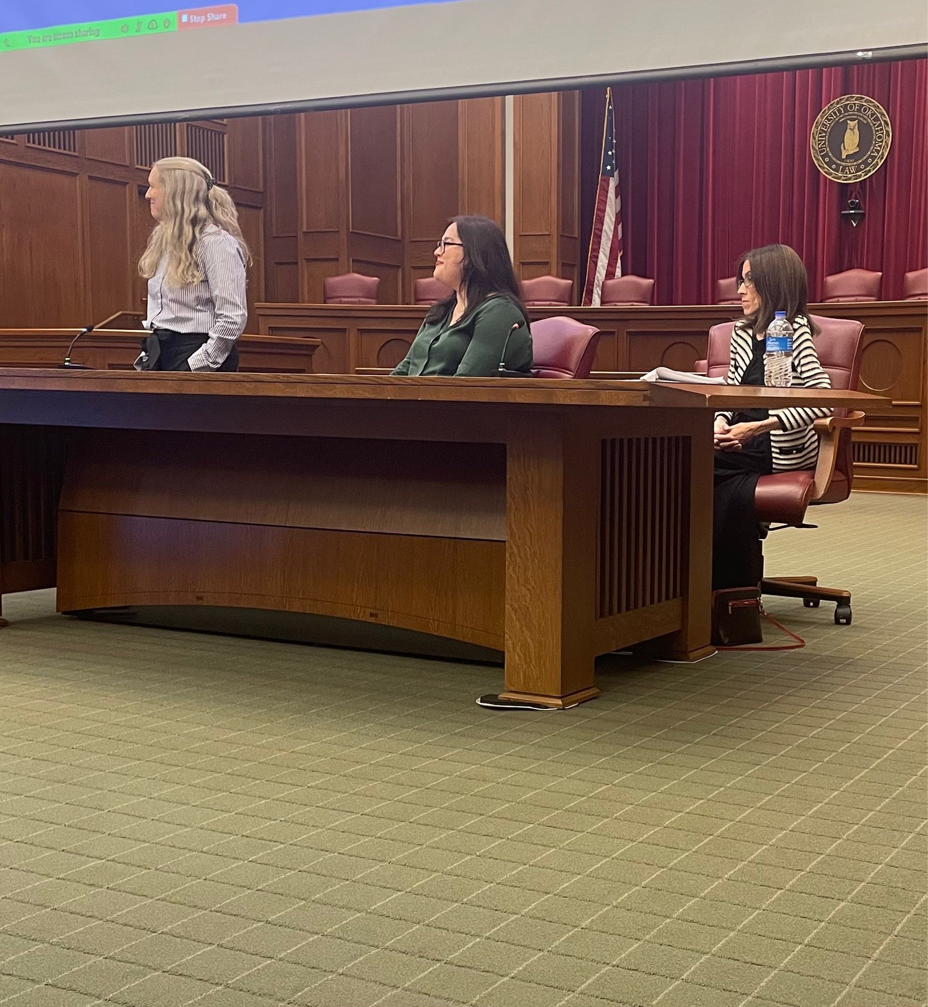 Three women sit at a table. One has long blonde hair, is wearing a blue striped shirt, and is looking to the right. One has long brown hair and is wearing a green shirt, and one is wearing a striped suit jacket with brown hair.