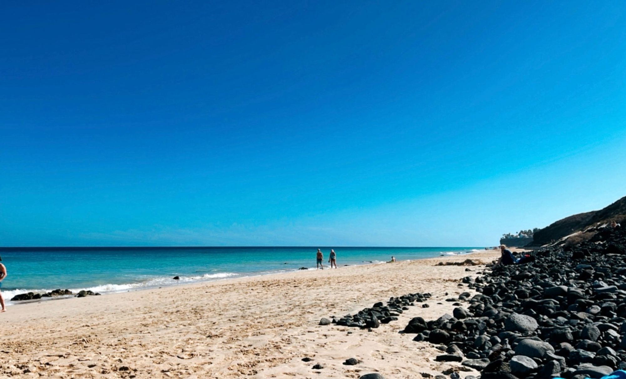 Langer Strand mit türkisblauen Wasser, gelben Sand und schwarzen Lavasteinen auf Fuerteventura mit 3 Menschen.