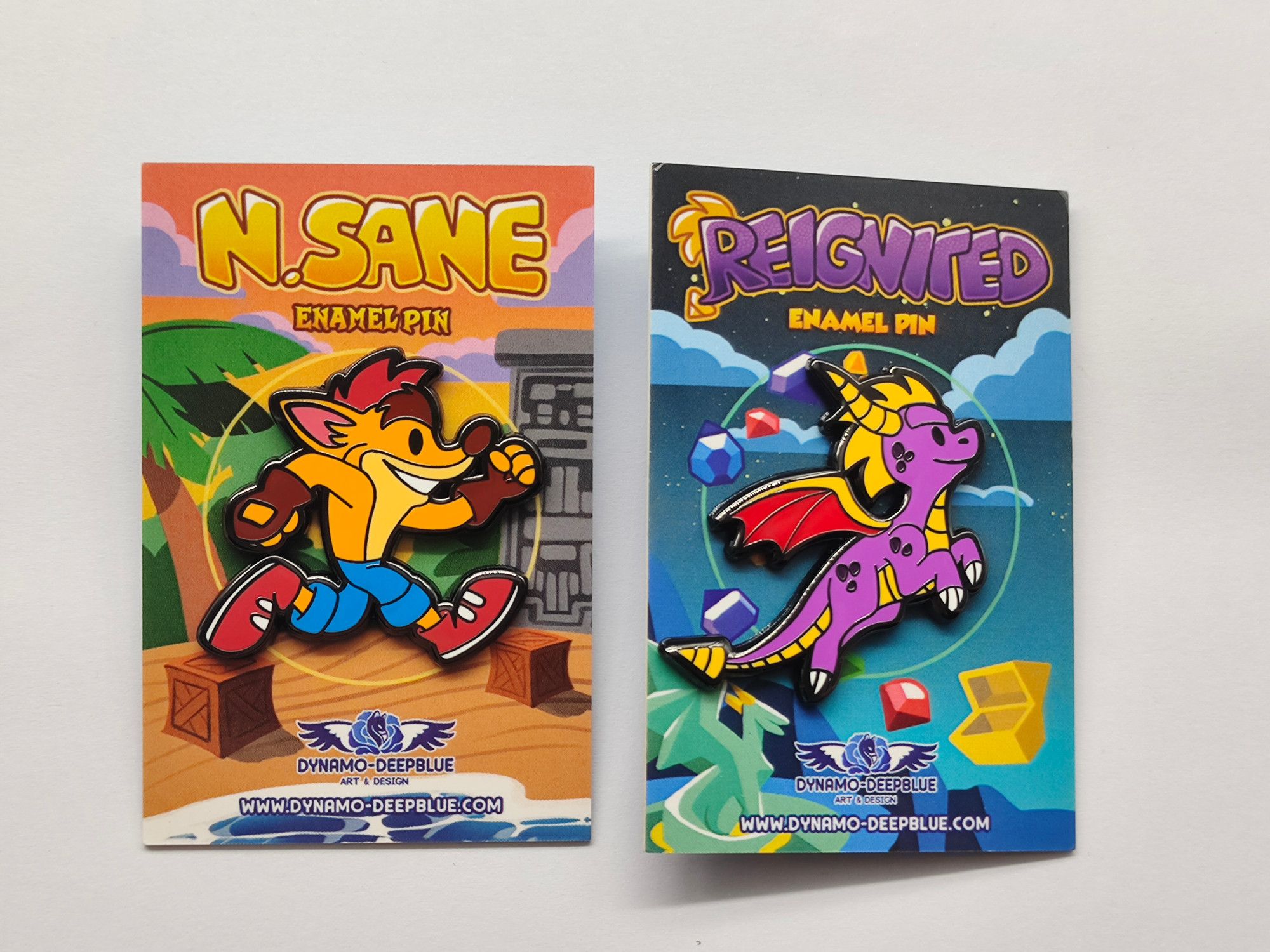 Photograph of two hard enamel pins, depicting a chibi Crash Bandicoot and a chibi Spyro the Dragon. They are placed on backing cards with illustrations themed to each franchise. The Crash pin's card reads "N. Sane enamel pin" at the top and the Spyro pin's card reads "Reignited Enamel Pin" at the top.