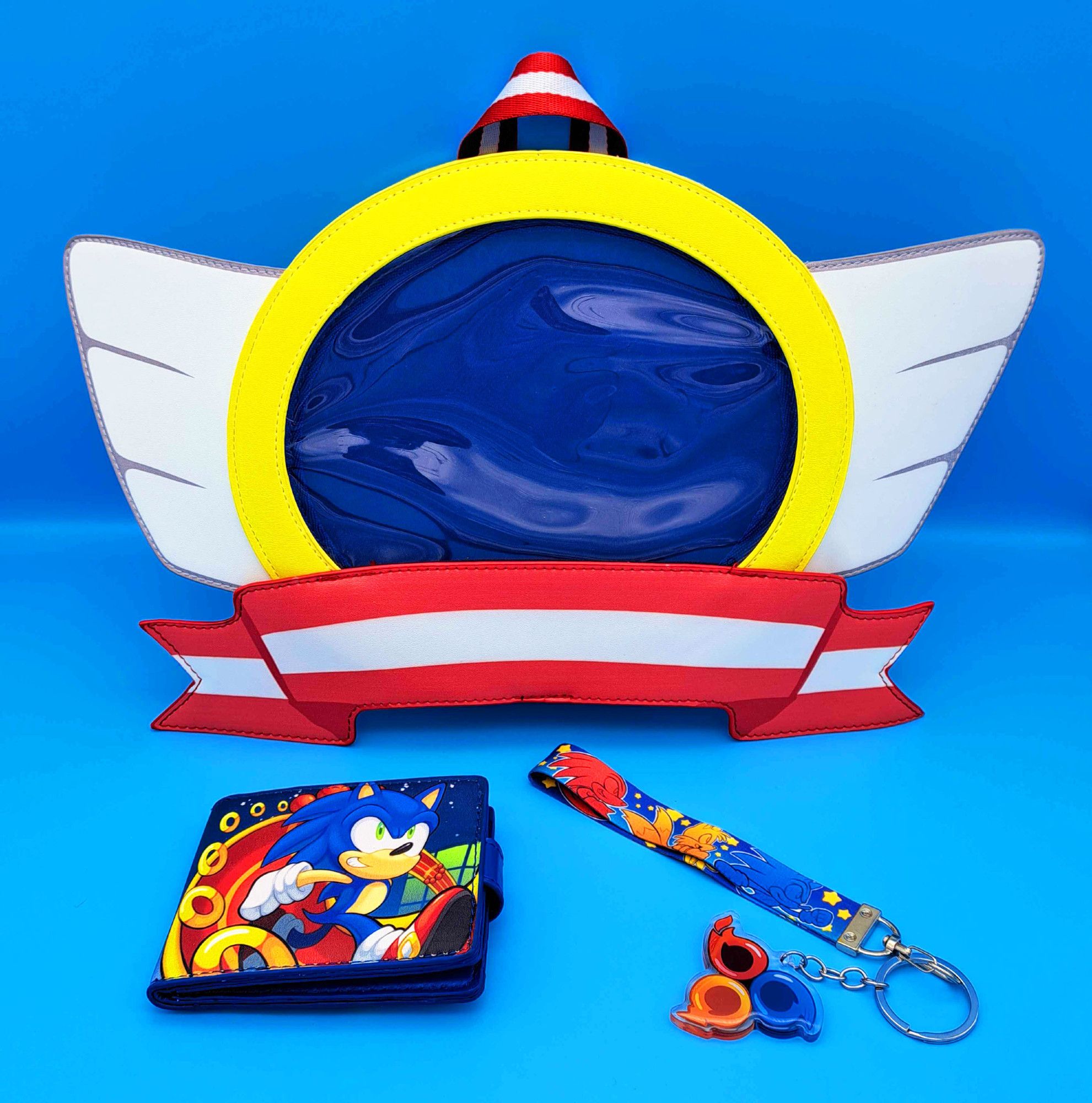 A photograph of an ita bag shaped like the emblems on title screens of various classic Sonic the Hedgehog games, a wallet with an illustration of Sonic on a special stage, and a wrist lanyard with an illustration of Sonic, Tails and Knuckles along with an attached keychain resembling the Sonic Heroes team emblem, against a blue backdrop.