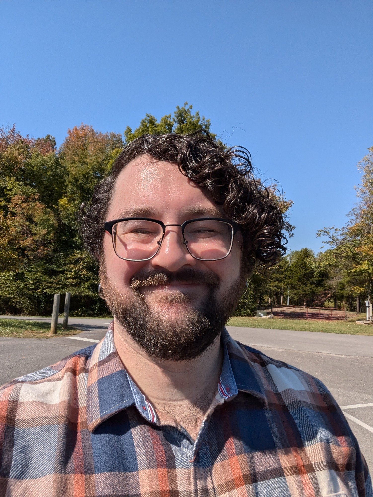 Male presenting person in an orange and blue flannel shirt standing in a parking lot with trees in the background. The trees are starting to turn from green to orange. The person has a close trimmed beard and dark curly hair that extends down to the bottom of their ear lobe on one side. The curls are very tight