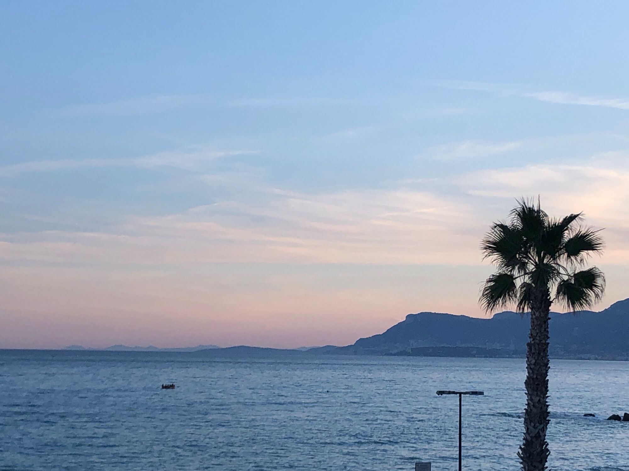 Blick auf das Meer und Berge im Hintergrund hinter denen die Sonne untergeht. Rechts im Vordergrund eine Palme.