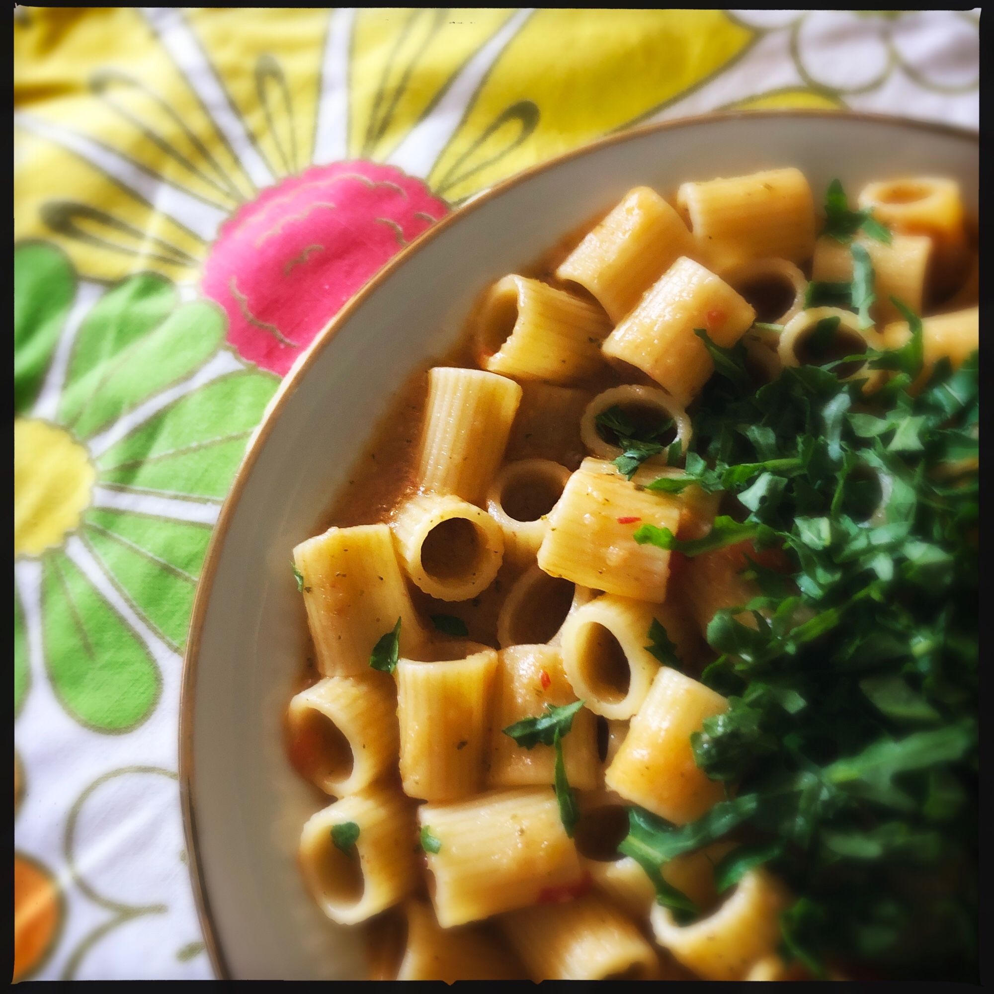 Ein weißer Teller Pasta mit Rucola bestreut. 

Auf der weißen Tischdecke sind große orangene und grüngelbe Blumen.