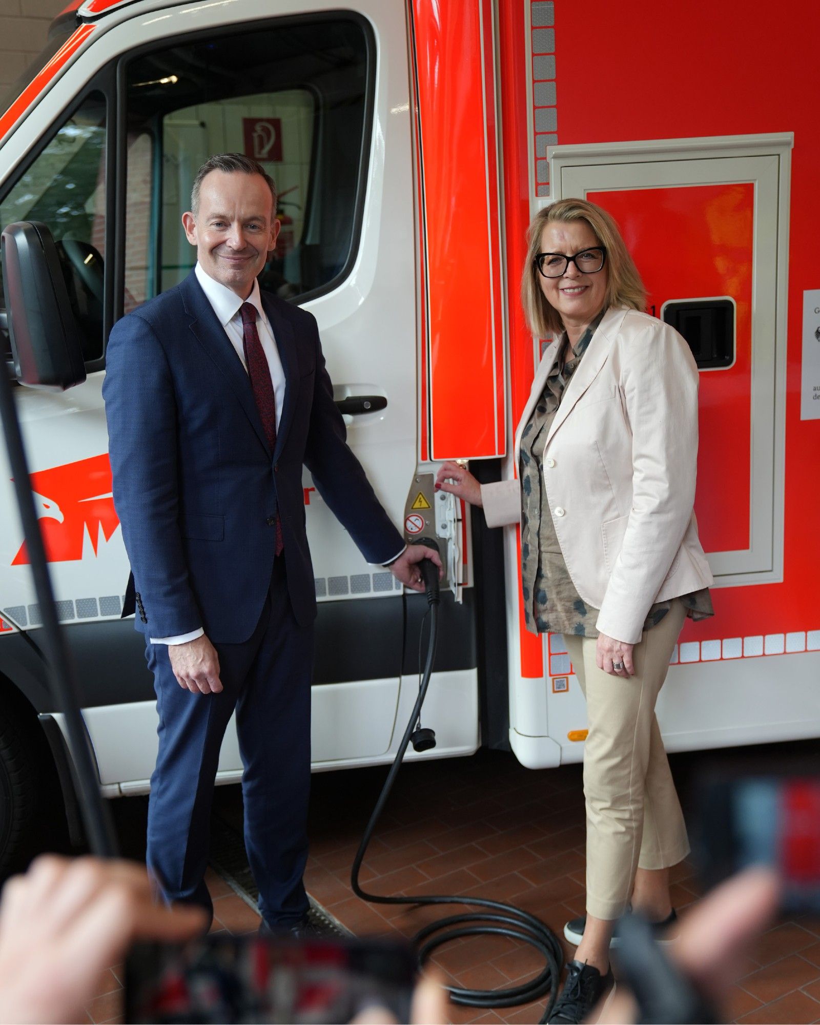 Stadtdirektorin Andrea Blome und Bundesverkehrsminister Dr. Volker Wissing stehen vor einem elektrisch angetriebenen Rettungswagen. Zwischen ihnen ist ein E-Ladekabel zu sehen, das in der Ladebüchse des Fahrzeugs steckt.