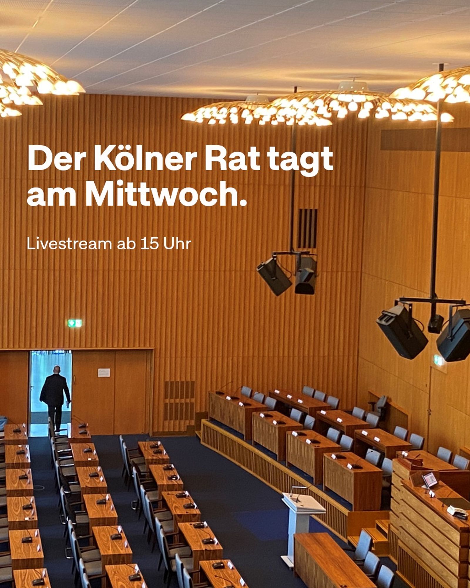 „Der Kölner Rat tagt am Mittwoch. Livestream ab 15 Uhr" steht in weißer Schrift auf einem Bild, das den Ratssaal von der Zuschauertribüne aus zeigt. Stühle, Tische und das Rednerpult sind nicht besetzt. Ein Mensch verlässt den Saal durch die Tür.