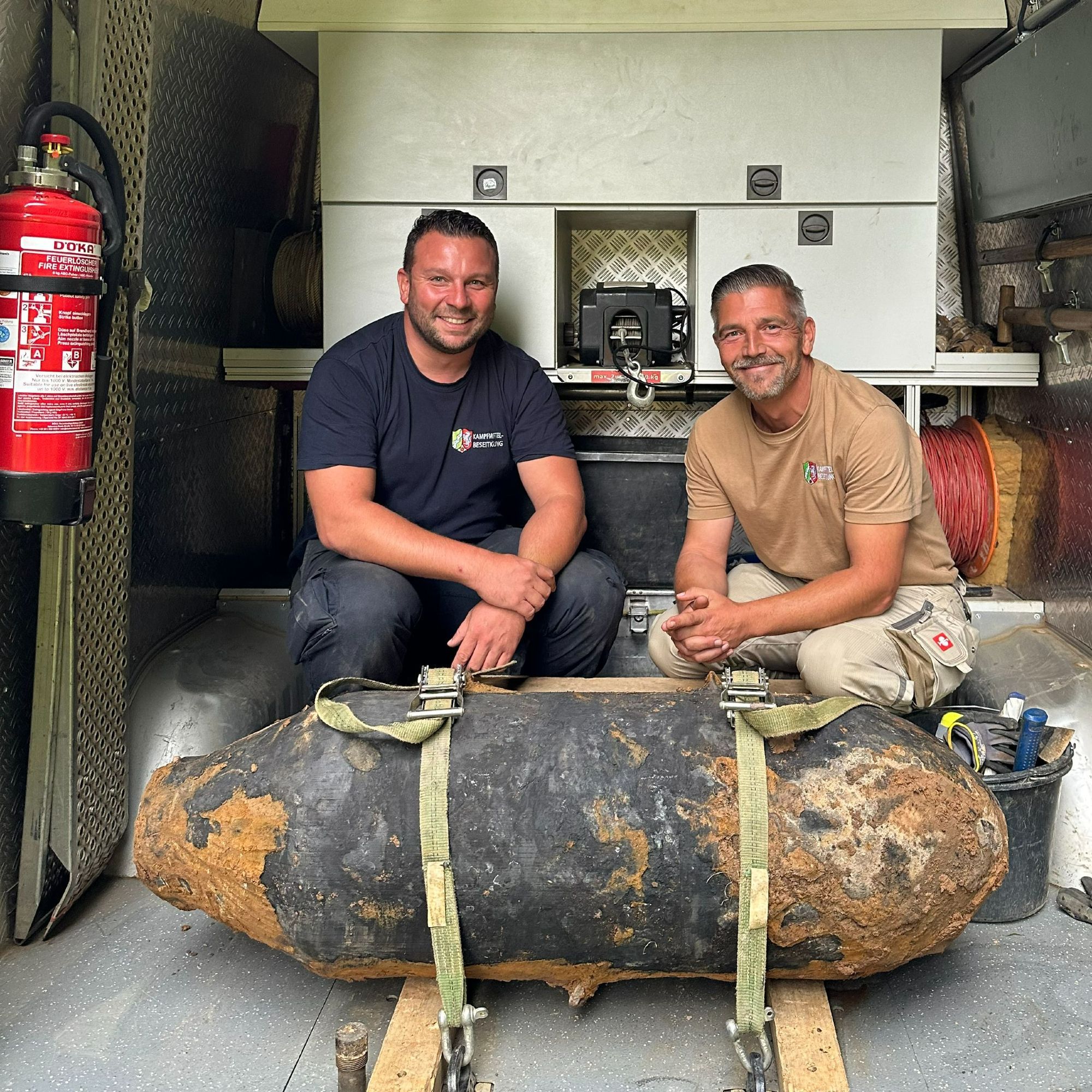 Auf dem Foto sind zwei Personen vom Kampfmittelbeseitungsdienst zu sehen. Sie hocken hinter einem Bombenblindgänger auf der Ladefläche eines Transporters.