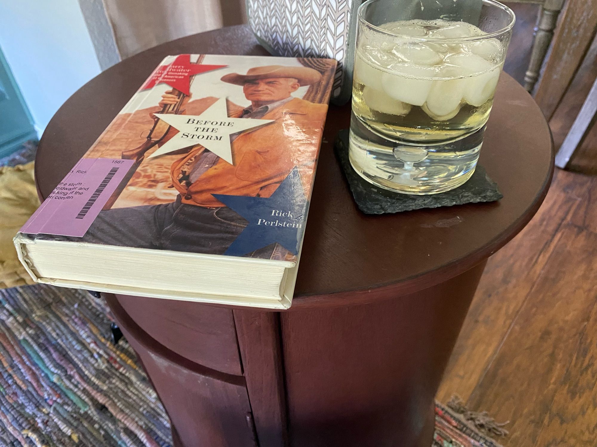 Before the Storm by Rick Perlstein, sitting on a side table next to a mixed whiskey drink in a rocks glass.