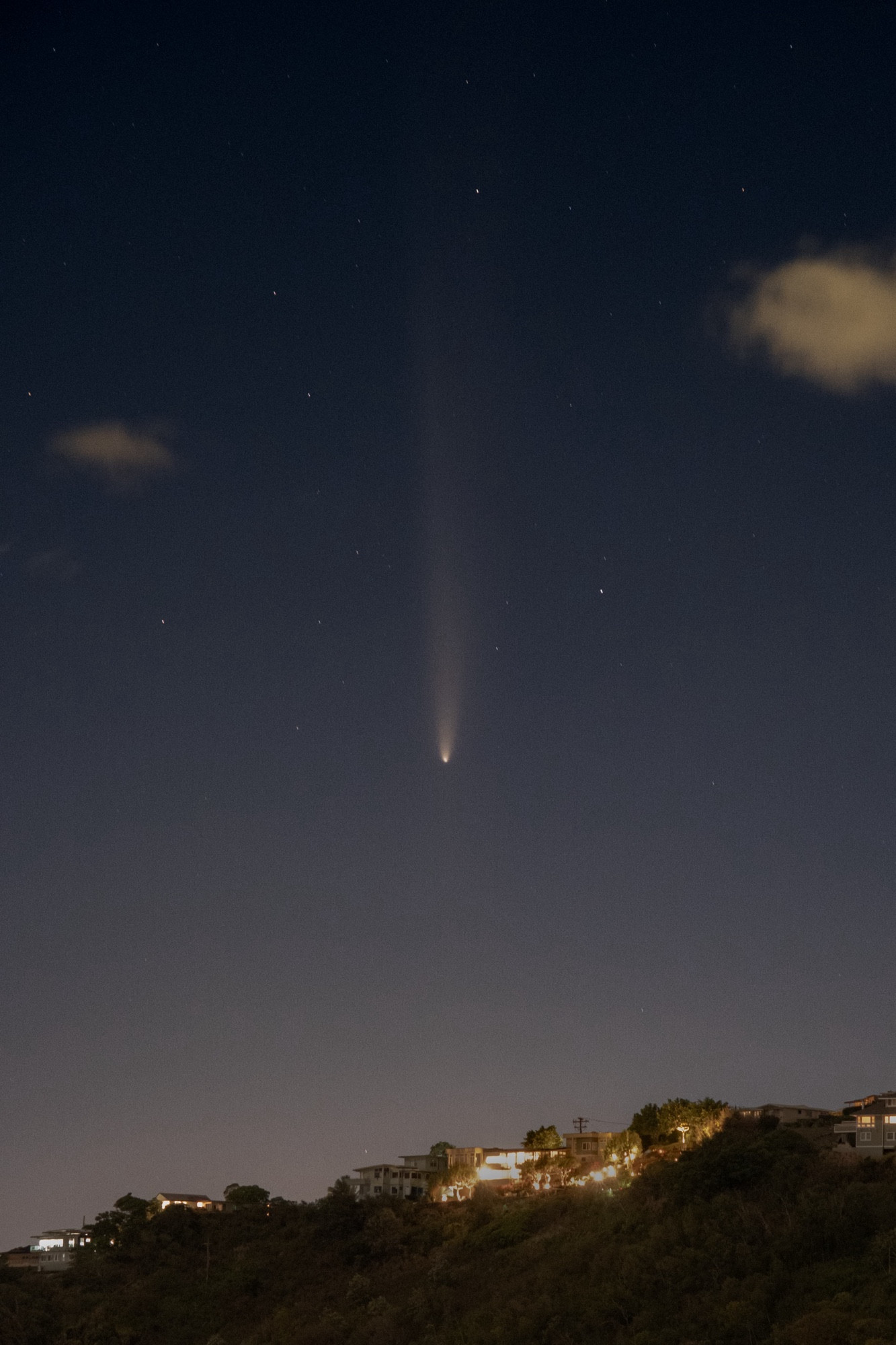 A comet descends vertically across the sky above a ridge with houses on it. 