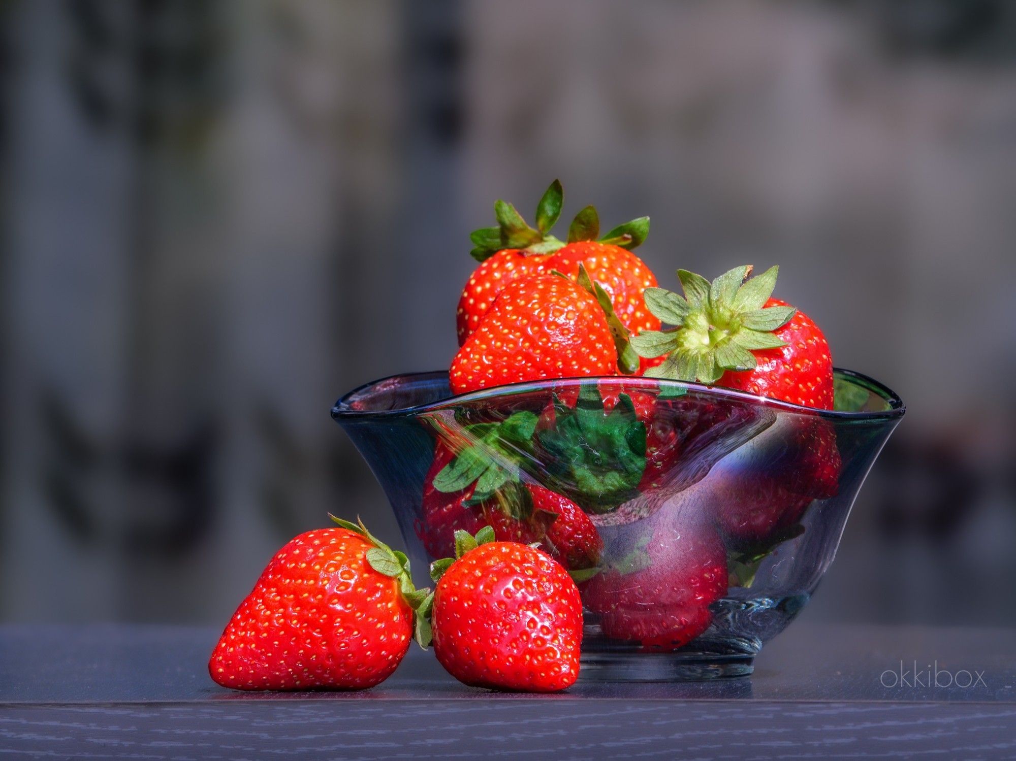 Aardbeien in een glazen schaaltje op tafel in de zon. Er liggen twee aardbeien naast het schaaltje.