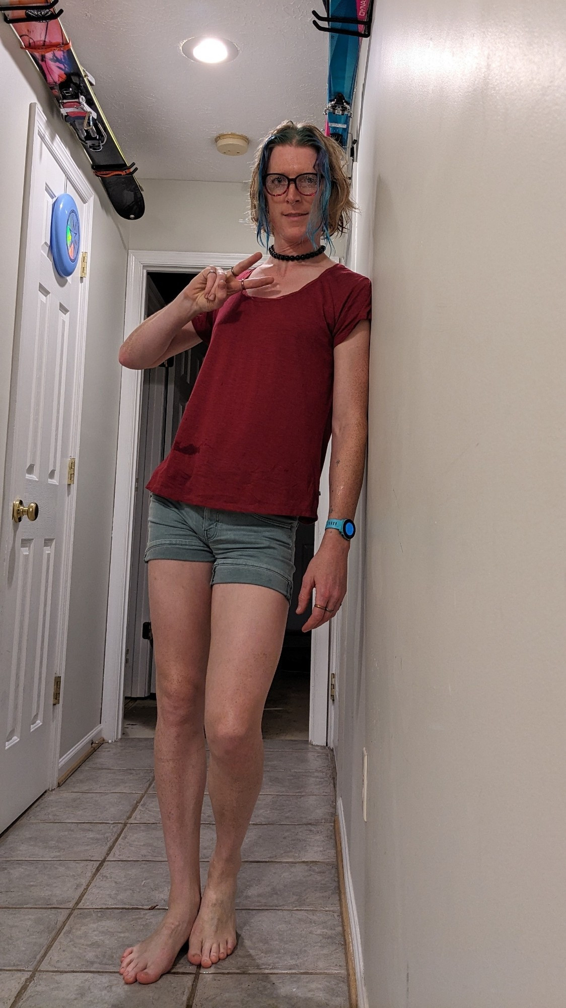 the same tall aquamarine and blonde haired lass standing in her hallway after taking all the rain gear off, her hair is wet but she and her clothes are dry. she is wearing a maroon scoop neck t-shirt and short green shorts.