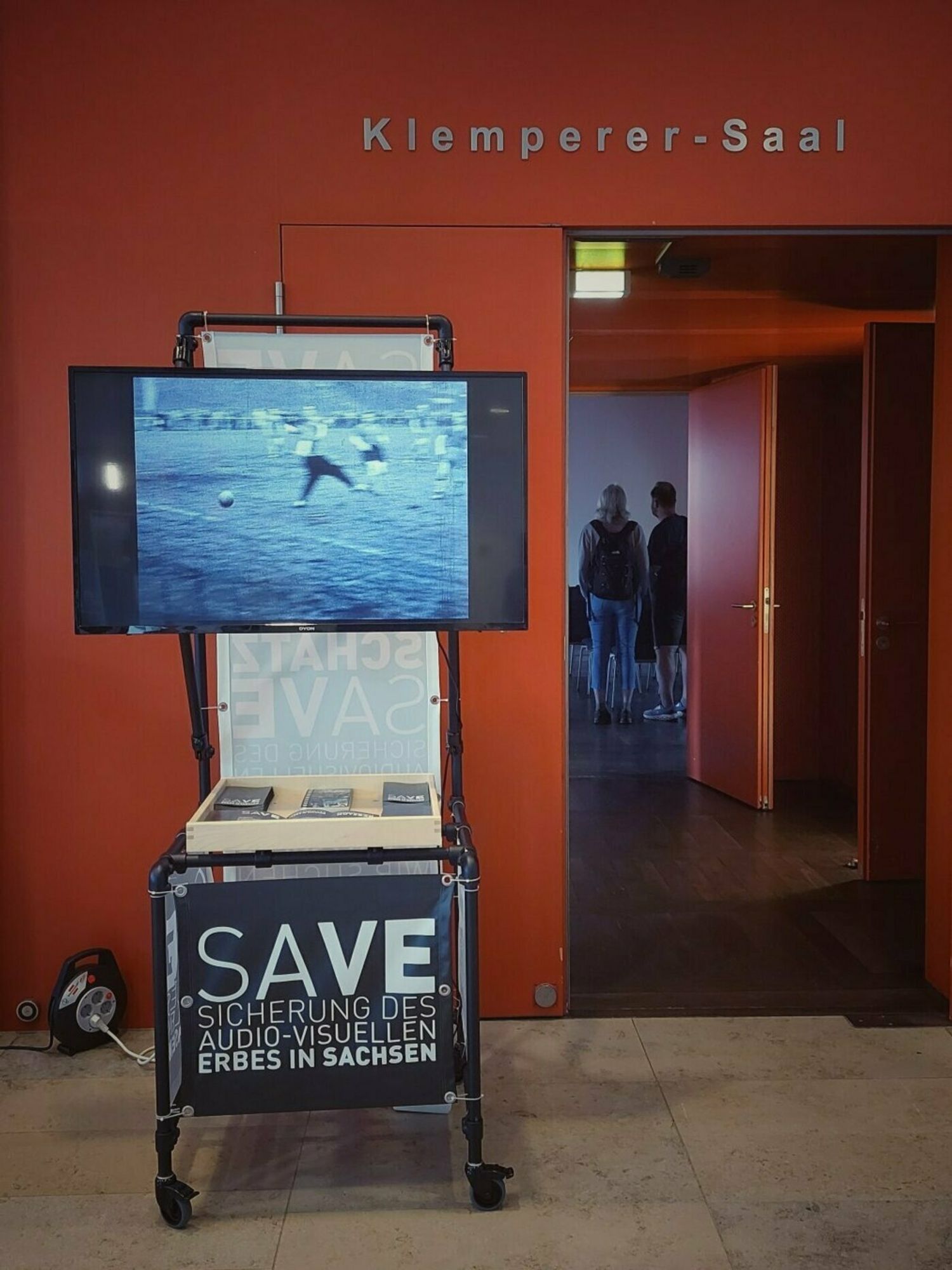 Türe zu einem Hörsaal, davor ein Monitor und ein Poster mit der Aufschrift SAVE.